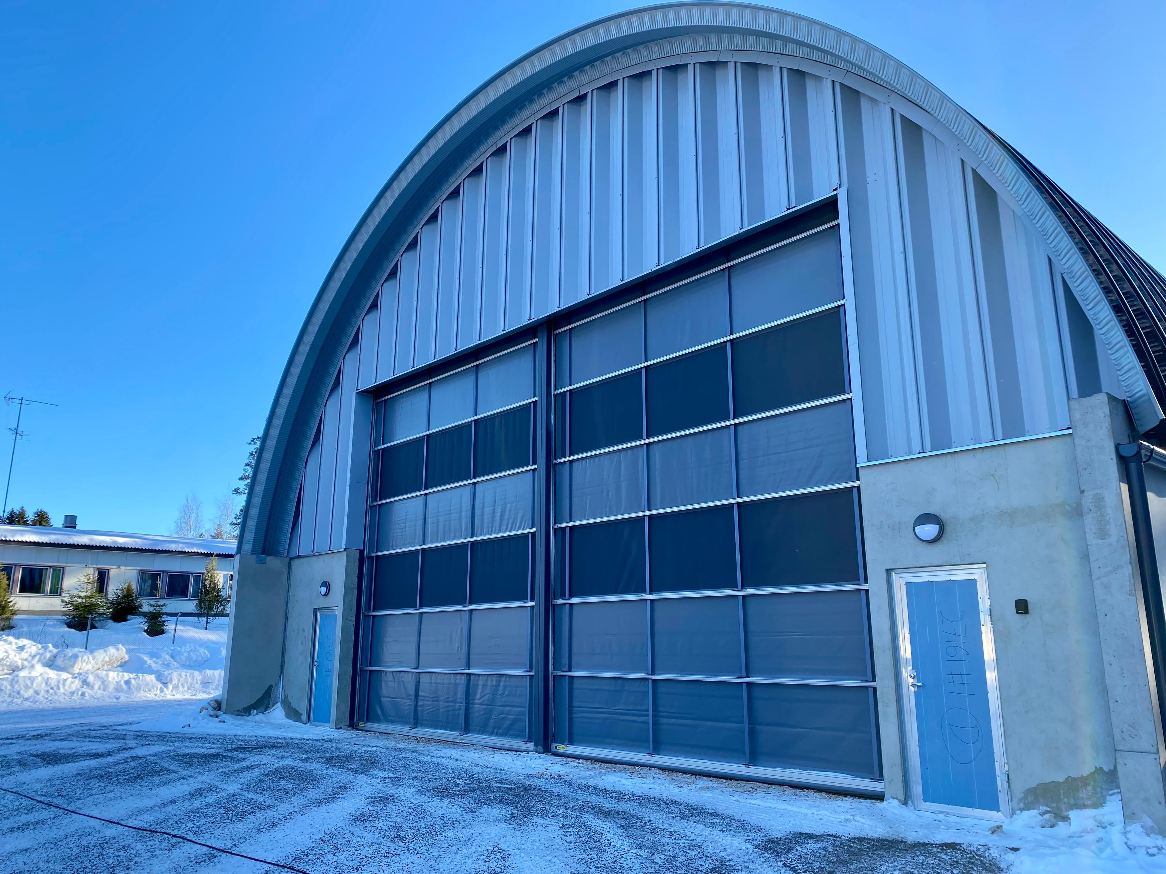 Kaaren muotoinen ja sinisävyinen rakennus, jossa isot liki kattoon asti avautuvat ovet. Talvisen maiseman taustalla näkyy kirkkaan sininen taivas. 