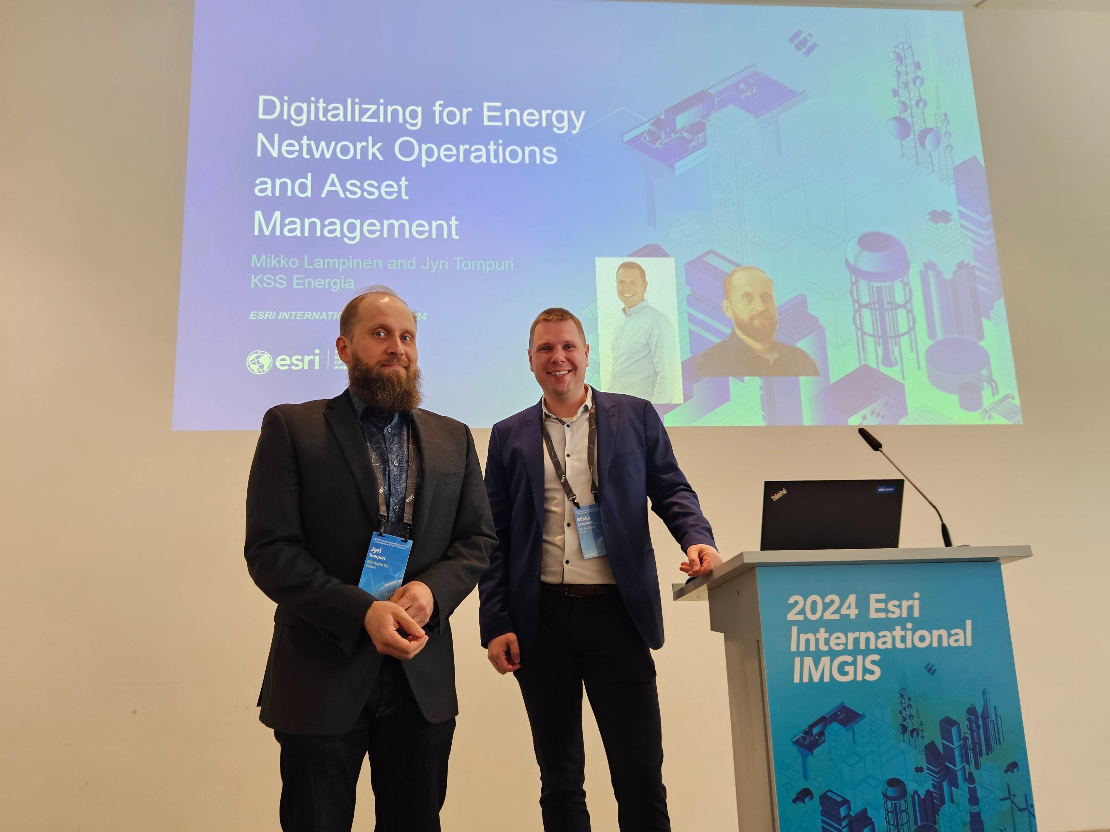 Kaksi pukuun pukeutunutta miestä katsoo hymyillen kameraan konferenssin lavalla. Taustalla näkyy seinälle heijastettu esitys sinivärinen esitys. Puhujapöntössä näkyy 2024 Esri International IMGIS -teksti.