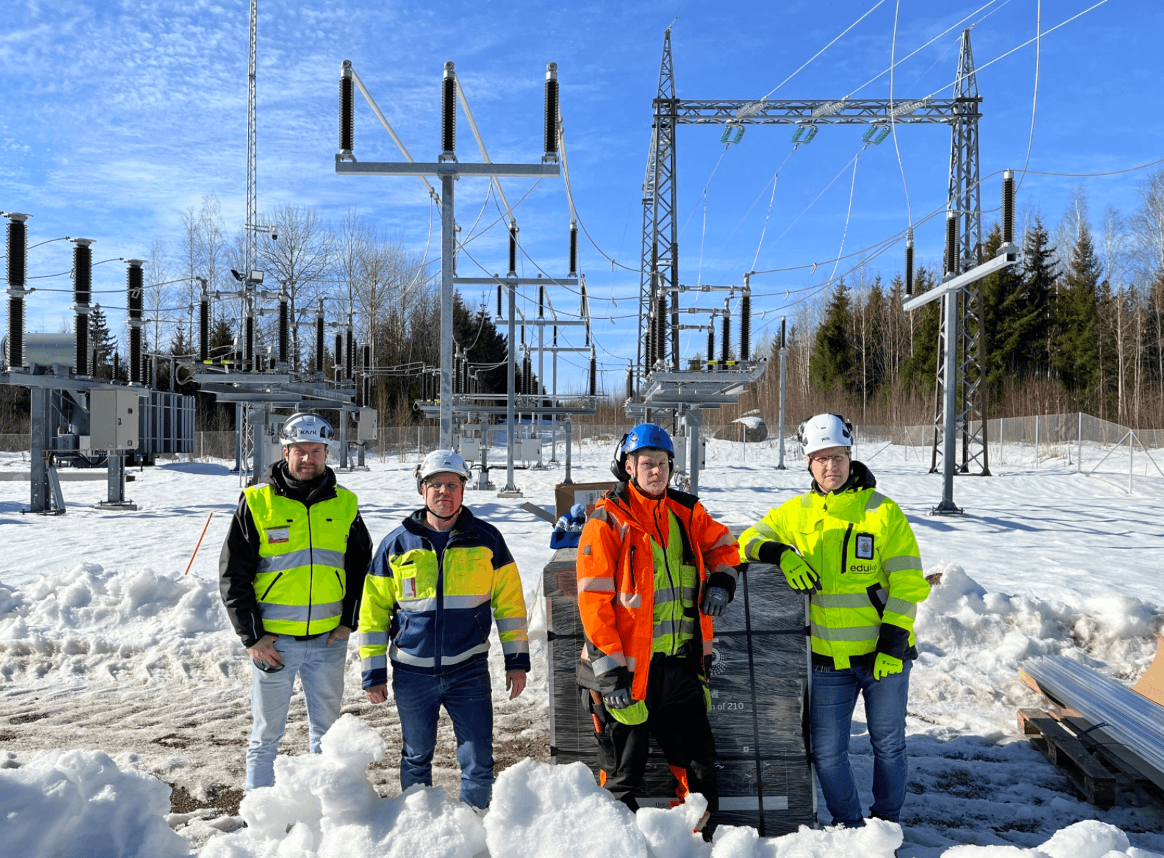 Neljä työmaavaatetukseen ja työmaakypäriin pukeutunutta miestä seisoo sähköaseman sähkölinjojen edustalla. Taustalla näkyy kirkas sininen taivas, maisema on talvinen ja luminen.