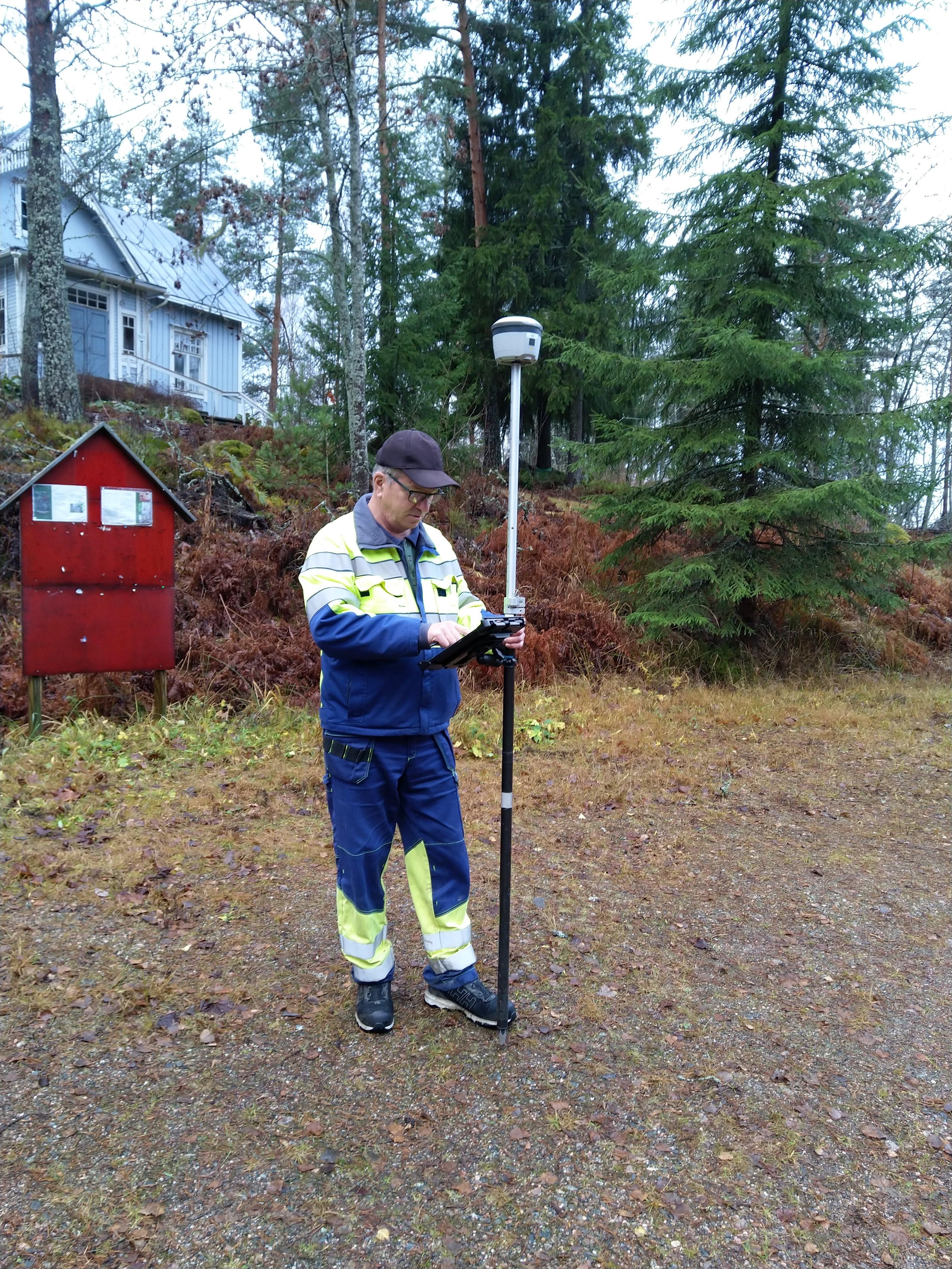 Henkilö työasussa GPS-mittasauva kädessään kirjaa tuloksia sauvassa kiinni olevaan tablettitietokoneeseen.