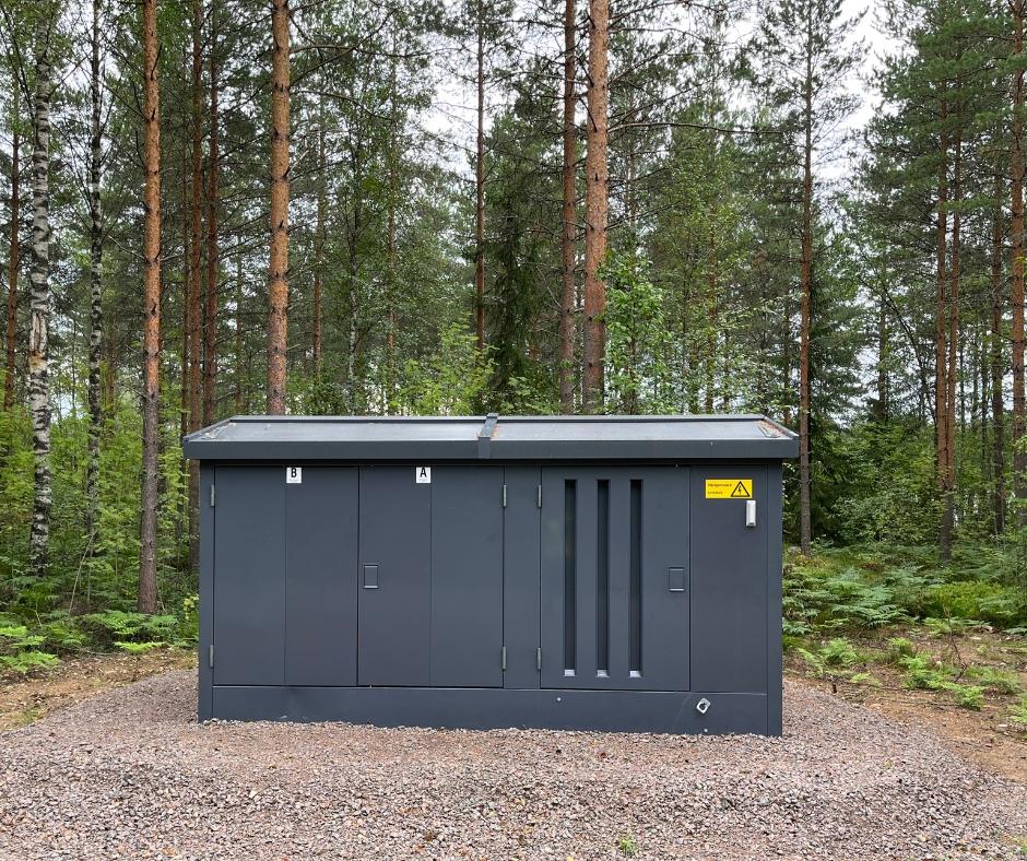 Tummanharmaa metallinen puistomuuntamokoppi havumetsämaisemassa.