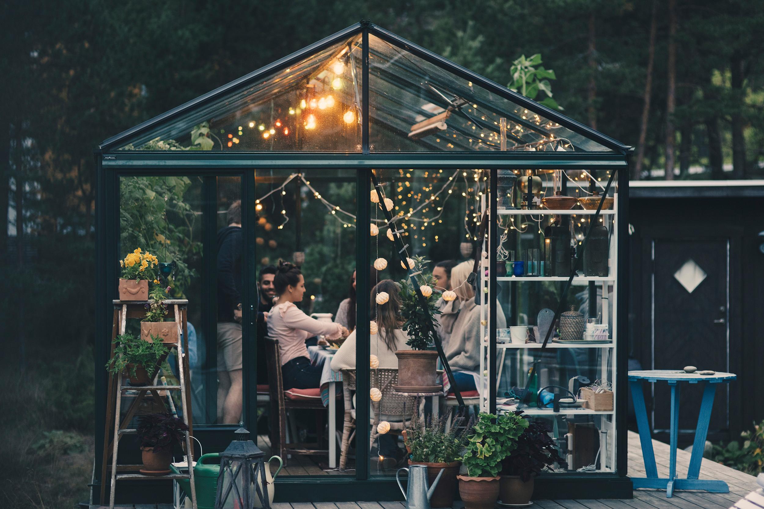 Lasisen kasvihuoneen sisällä on ihmisiä istumassa pöydän ääressä. Kattoon ripustetut valonauhat luovat tunnelmaa.