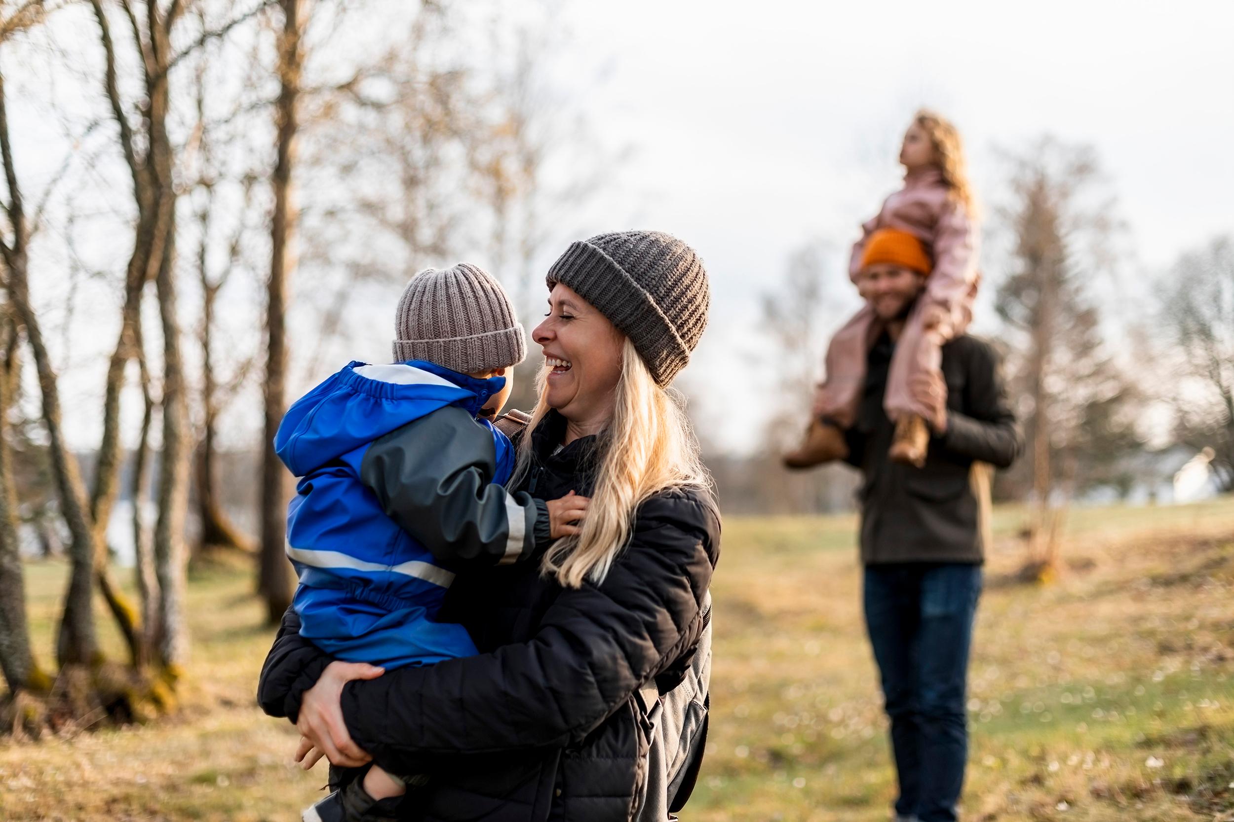 Perhe retkeilee ulkona syksyisessä maastossa. Puissa ei ole enää lehtiä. Vaalea hiuksinen nainen pitelee sylissään sinipukuista lasta. Taustalla mies pitelee olkapäillään vaaleanpuna pukuista lasta. 