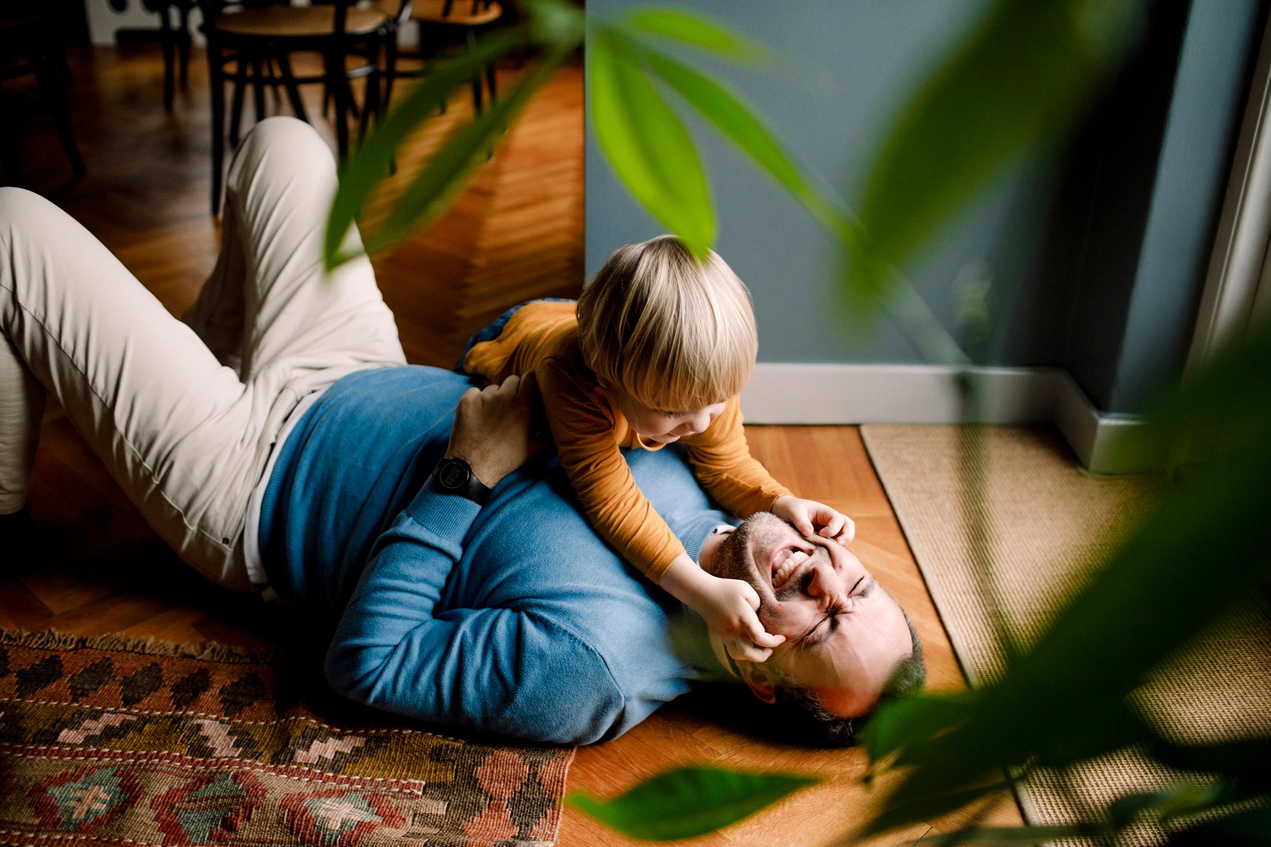 Sinipaitainen isä ja oranssipaitainen lapsi leikkivät kodin puisella lattialla. Leikki-ikäinen lapsi puristaa käsillä nauravan isän kasvoja. Kuvan edustalla näkyy viherkasvin lehtiä.