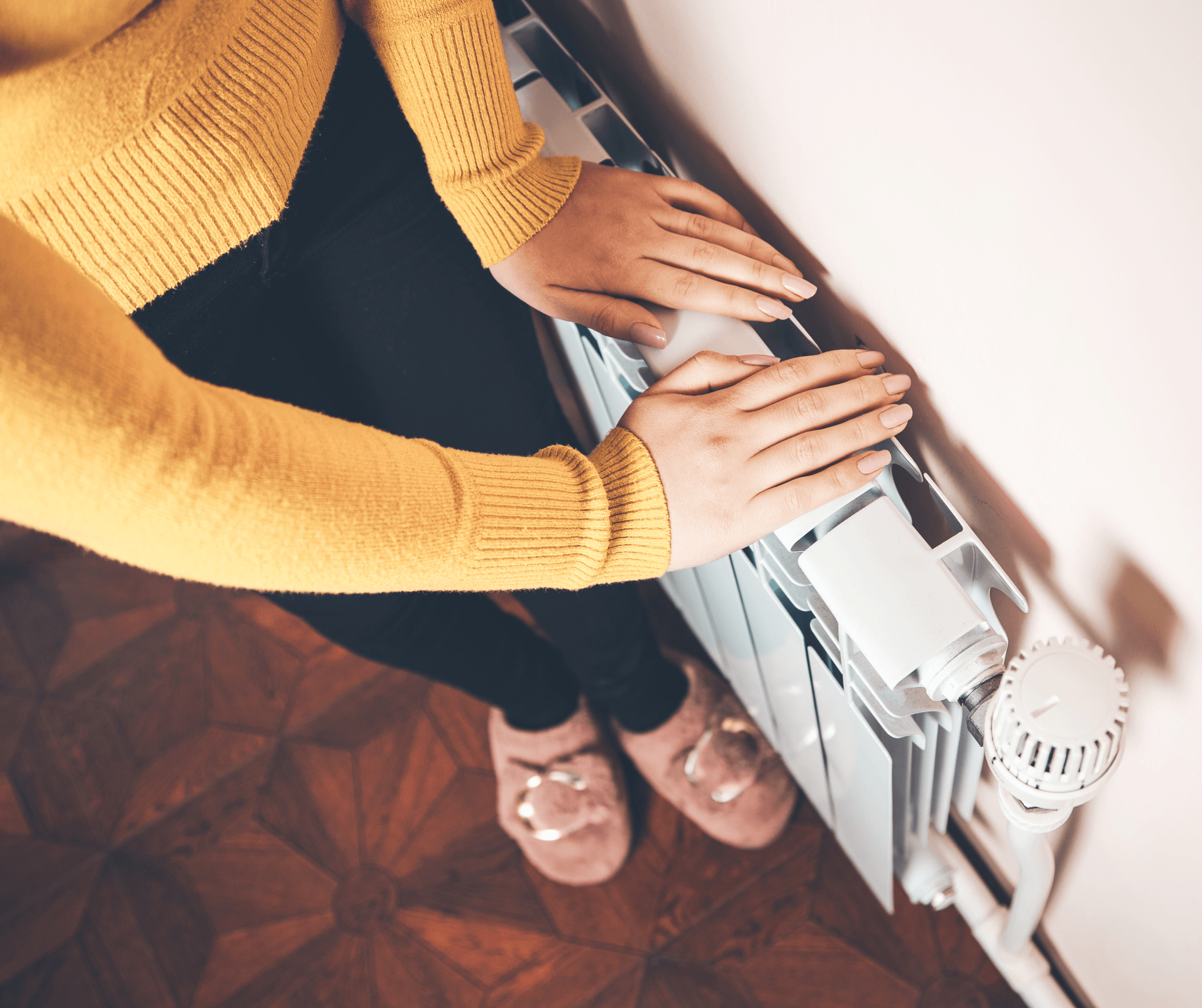 Keltapaitainen nainen seisoo seinustan vieressä molemmat kädet seinäpatterin päälle asetettuna, kokeilee pattereiden lämpöä.