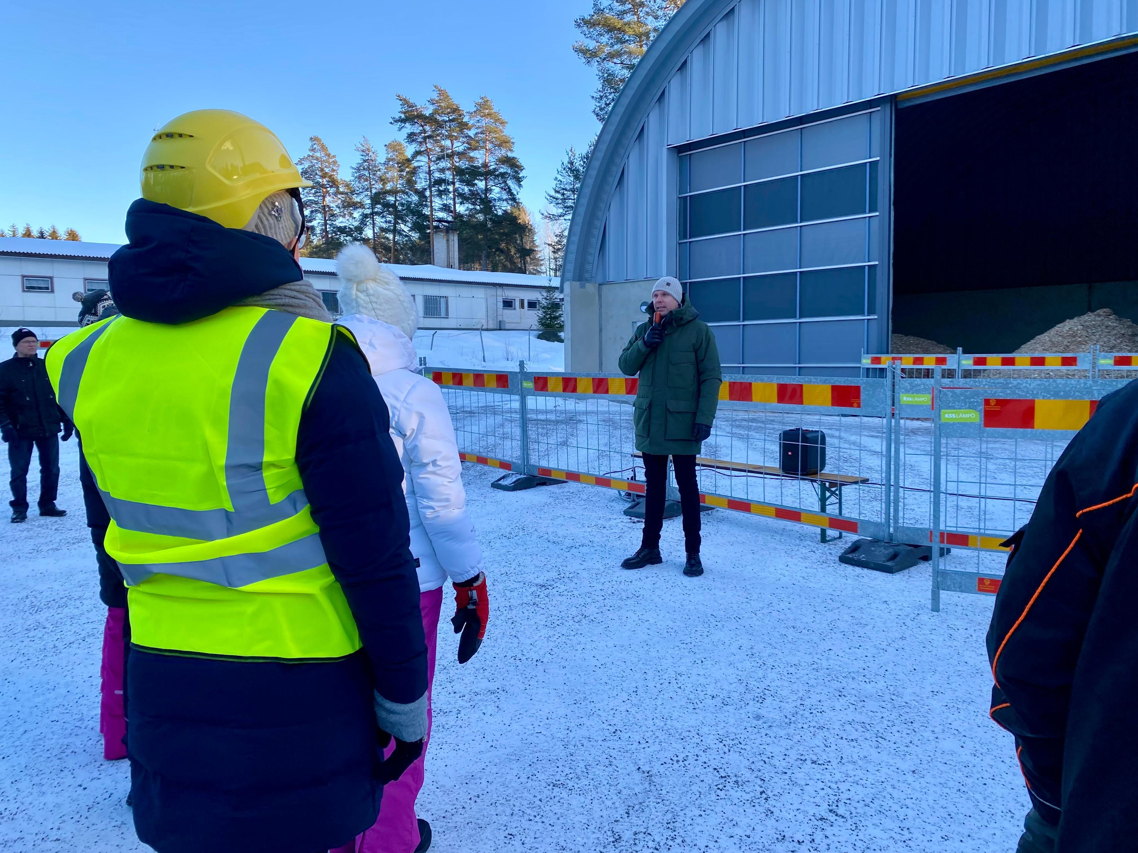 Vihreään talvitakkiin pukeutunut mies pitää puhetta biolämpölaitoksen edustalla. Kuvan etualalla on ihmisiä kuuntelemassa puhetta, yhdellä henkilöllä on huomioliivi ja keltainen kypärä. Puheenpitäjän taustalla on työmaa-aita, jonka takana näkyy vaaleansininen rakennus, hakesiilo.