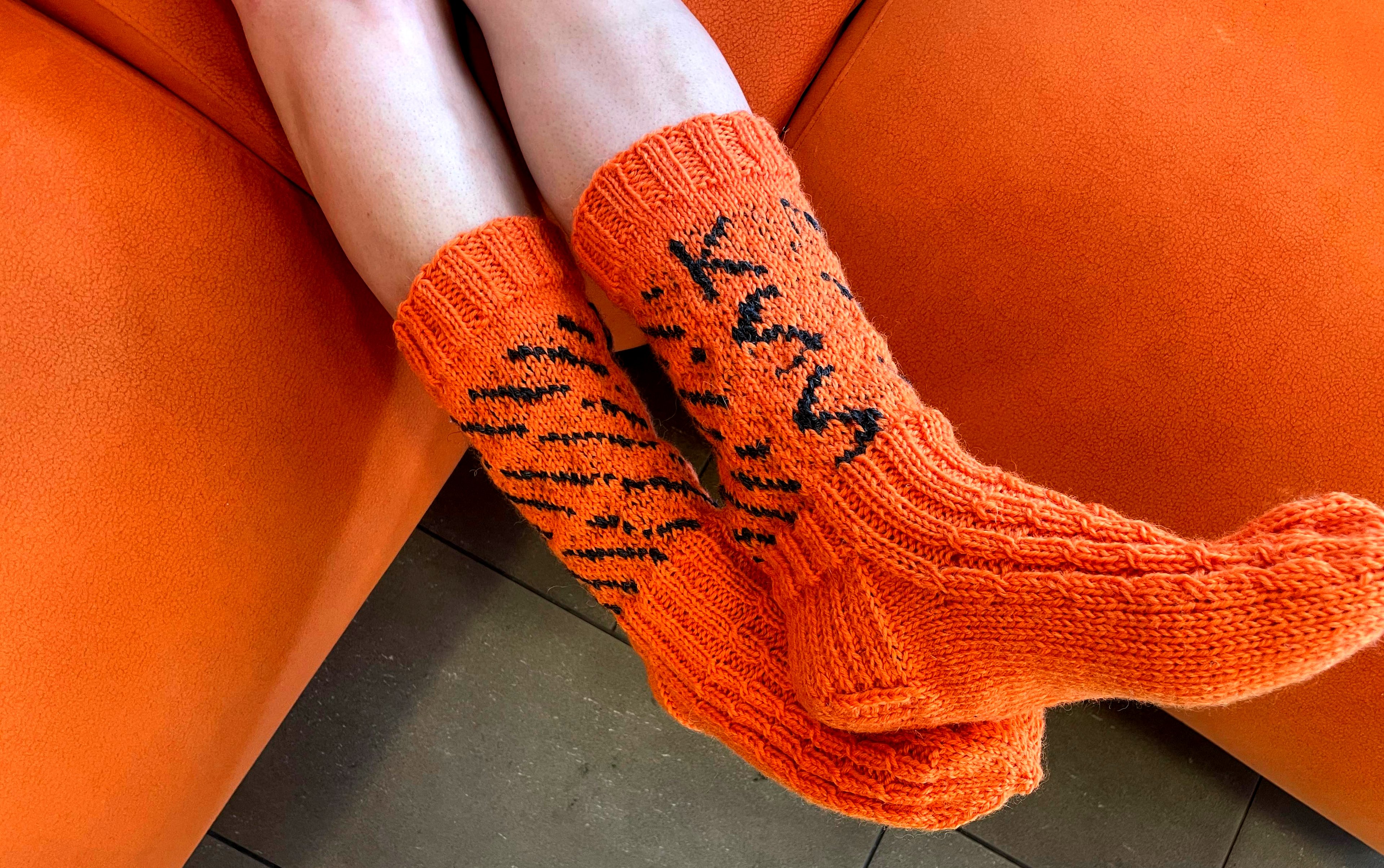 Oranssinväriset itse kudotut villasukat KSS Energia -brändin värisenä oranssina ja tekstinä sukkien varressa mustalla KSS. Sukissa on myös mustia viivoja koristeena.  Sukat ovat jalassa henkilöllä, joka istuu oranssilla sohvalla. Hänestä näkyy vain hieman jalkoja.