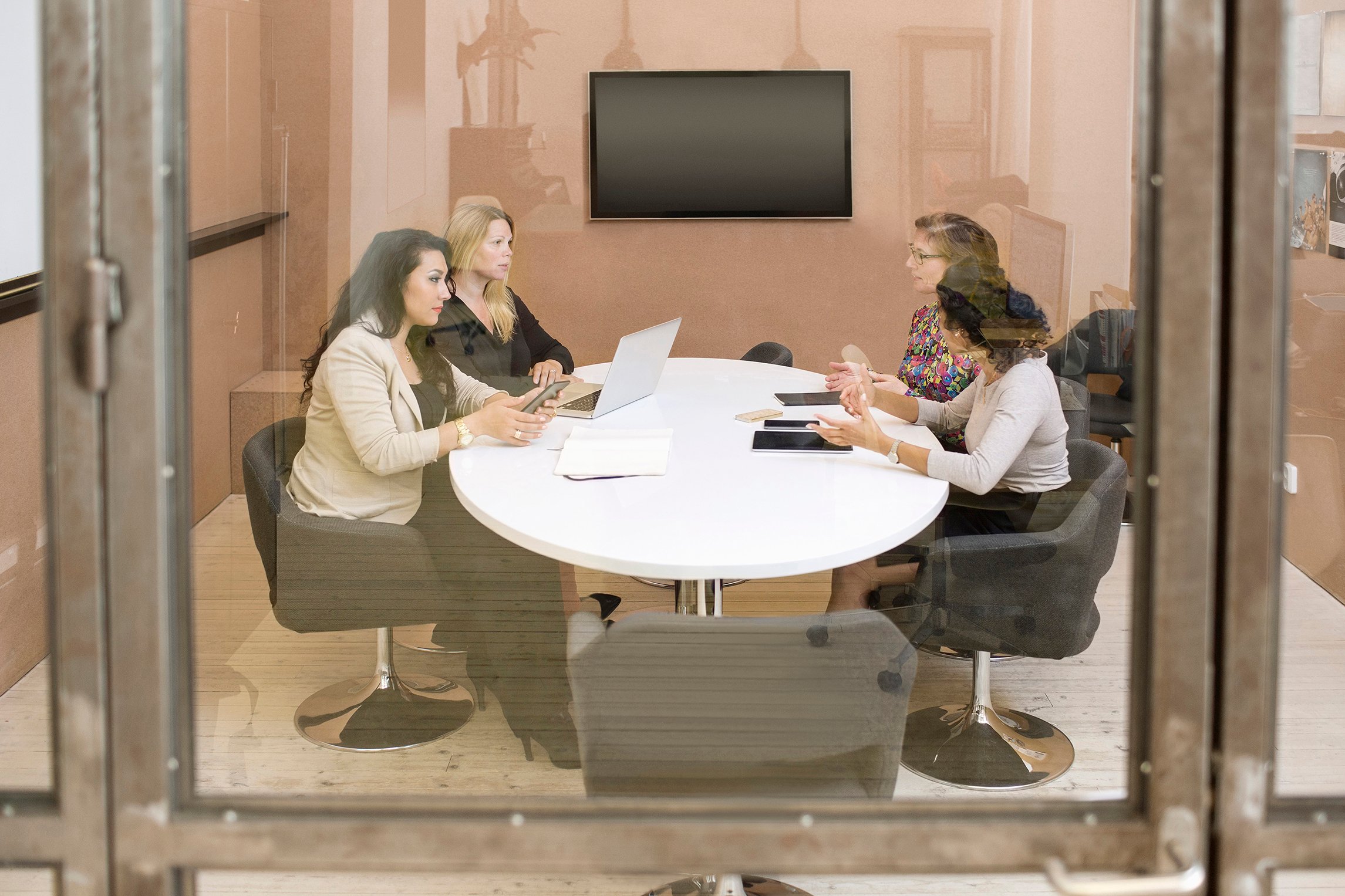 Neljä naishenkilöä istuu valkoisen pyöreän pöydän ääressä tummilla nojatuoleilla lasiseinäisessä toimistohuoneessa. Taustalla näkyy vaaleanpunainen seinä, jossa on musta kiinni oleva taulutelevisio. Naisten keskustelevat keskenään ja pöydällä on avoimena oleva kannettava tietokone sekä matkapuhelimia ja papereita.