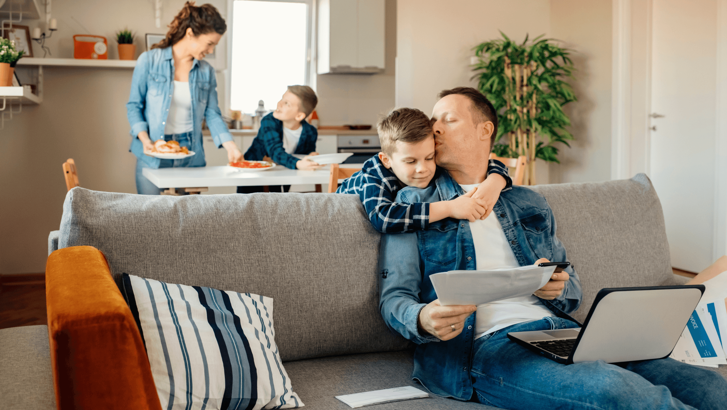 Perhe yhdessä kotona. Isä istuu sohvalla ja suukottaa ohimolle sohvan takana olevaa poikaa. Isän sylissä on kannettava tietokone ja käsissä papereita. Taustalla on äiti ja toinen poika kattamassa ruokapöytää.