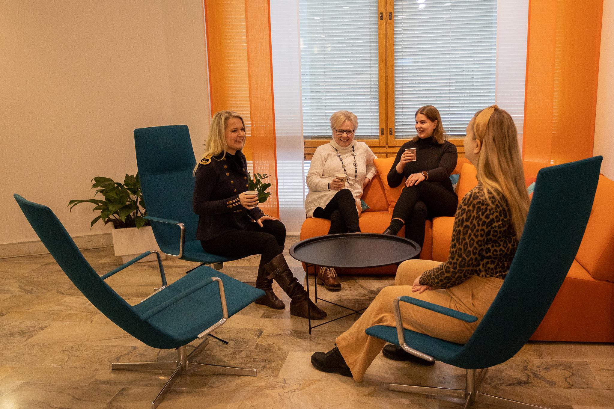 Neljä iloista naista istuu kahvilla. Kaksi naista istuu oranssilla sohvalla ja kaksi sinisissä nojatuoleissa. Taustalla on ikkuna, kasveja ja oranssit verhot. Kaikilla on kädessä kahvikupit. 