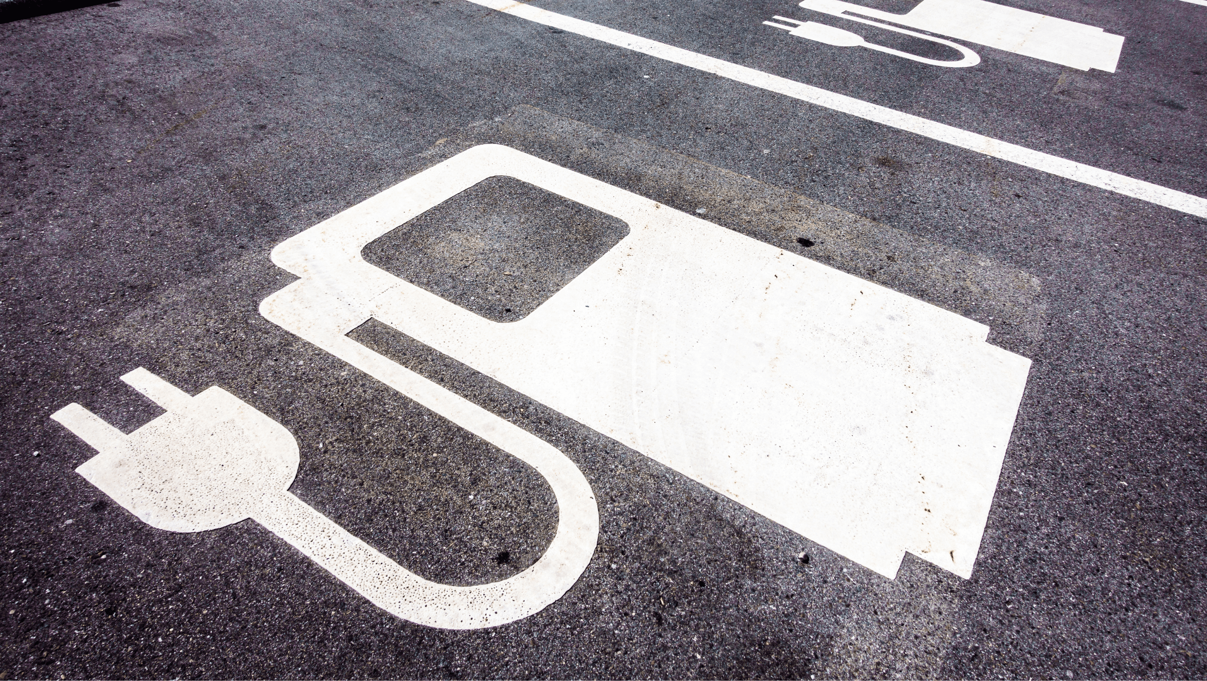 Harmaa asfaltoitu parkkiruutu, jossa valkoinen maalattu sähköautoilun latauspiste.