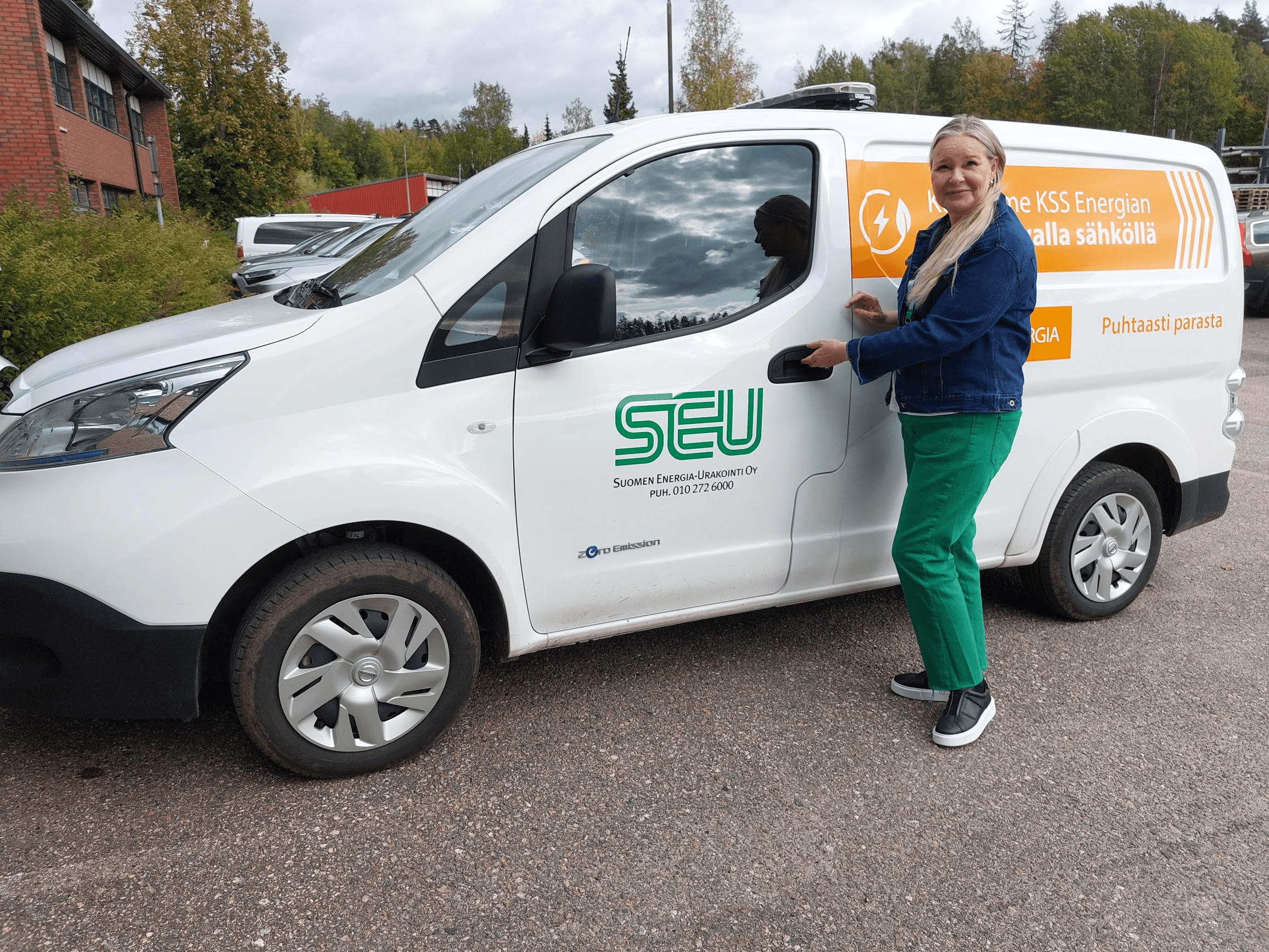 Suomen Energia-Urakoinnin myyntipäällikkö Marja Karvonen seisoo SEU:n valkoisen pakettiauton edustalla ja katsoo kohti kameraa hymyillen. Taustalla näkyy punatiilinen rakennus ja puita. Vuodenaika on syksy. 