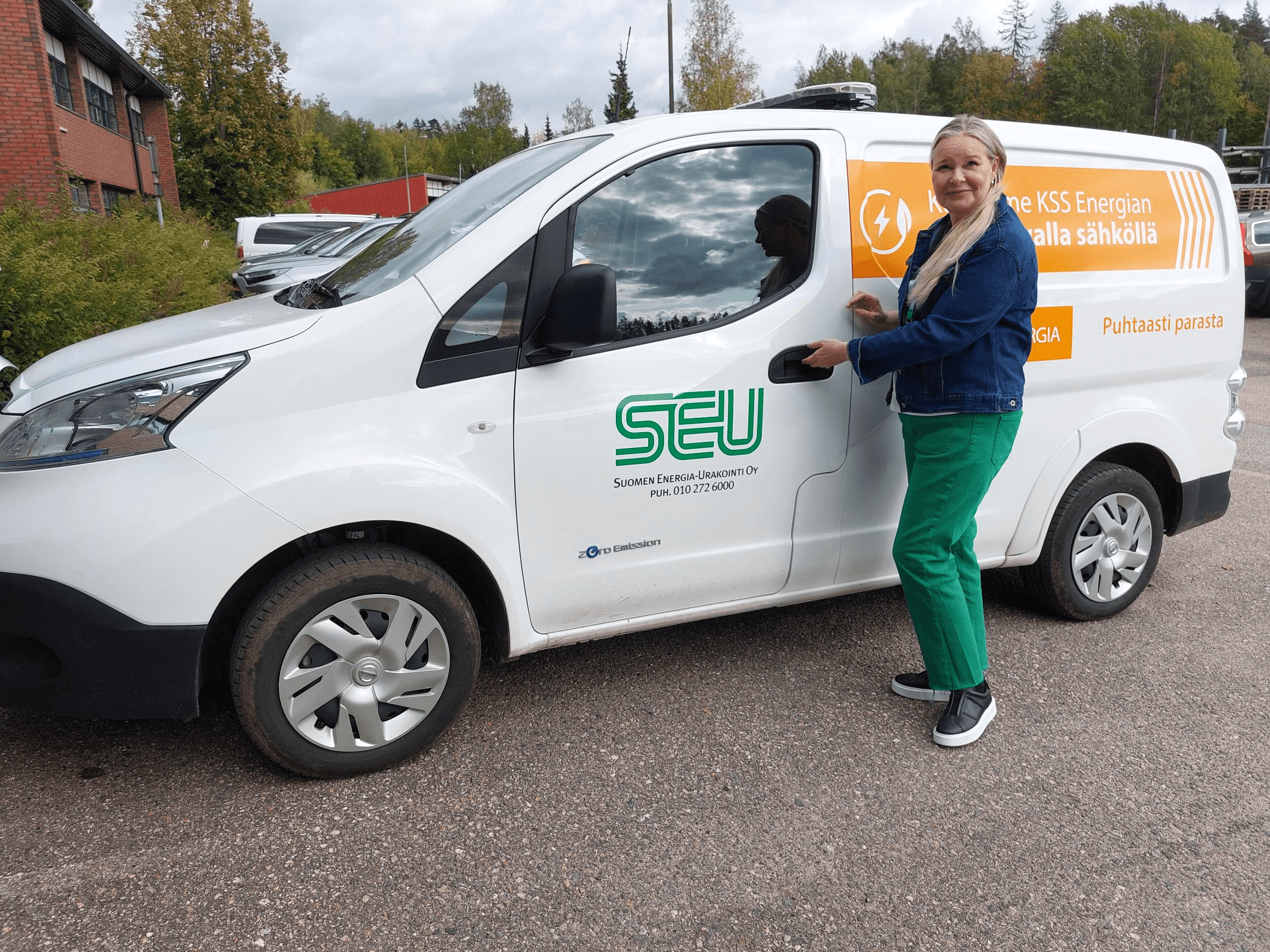 Suomen Energia-Urakoinnin myyntipäällikkö Marja Karvonen seisoo SEU:n valkoisen pakettiauton edustalla ja katsoo kohti kameraa hymyillen. Taustalla näkyy punatiilinen rakennus ja puita. Vuodenaika on syksy.