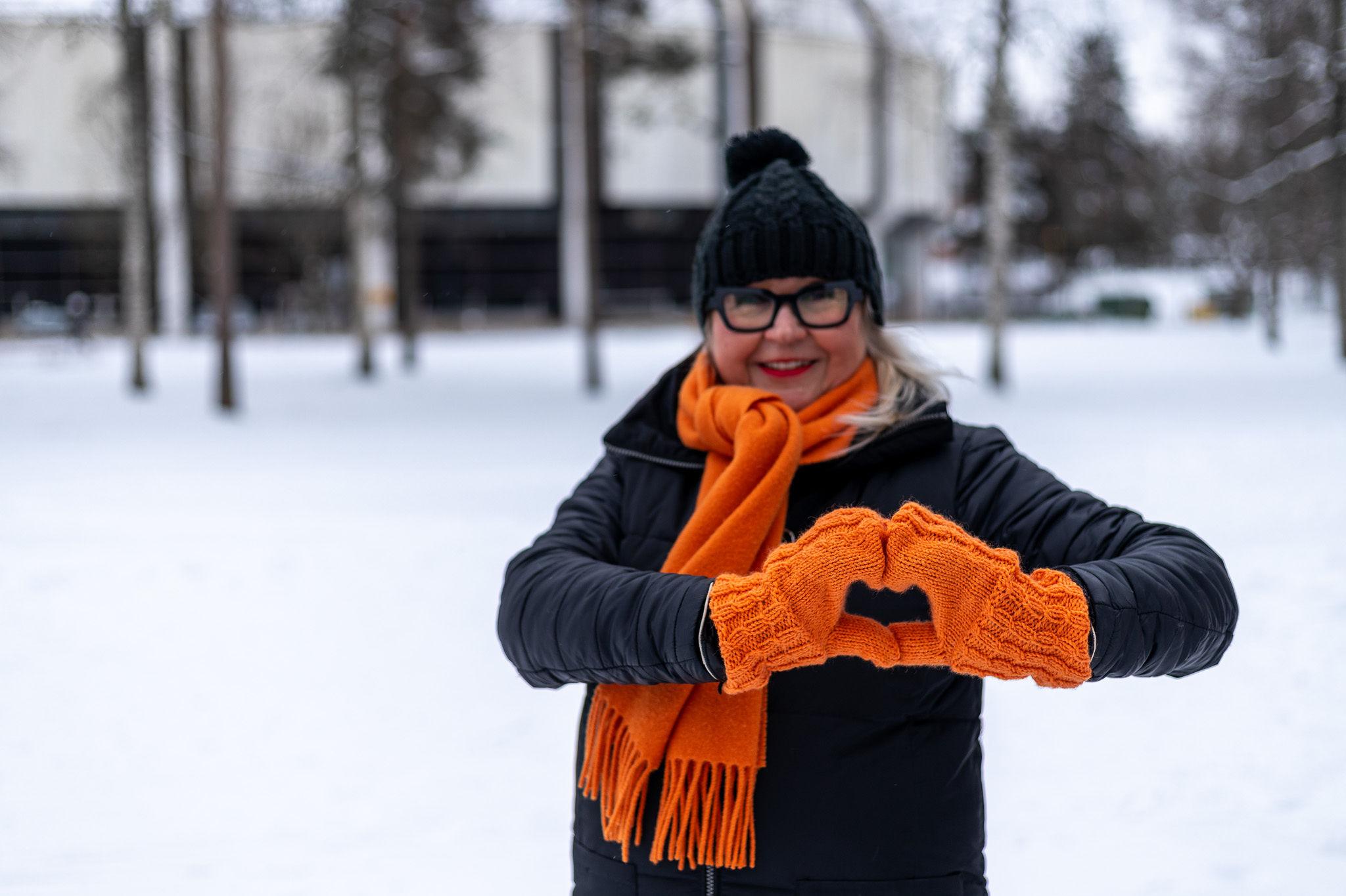 Nainen tekee käsistä sydämen ulkona talvella. Hänellä on kädessä oranssit lapaset ja kaulassa oranssi kaulaliina. Naisella on päässä musta pipo ja päällä musta toppatakki. Hänellä on mustat silmälasit ja hymyilevät punaiset huulet. Taustalla näkyy lunta ja puita sekä jäähalli.