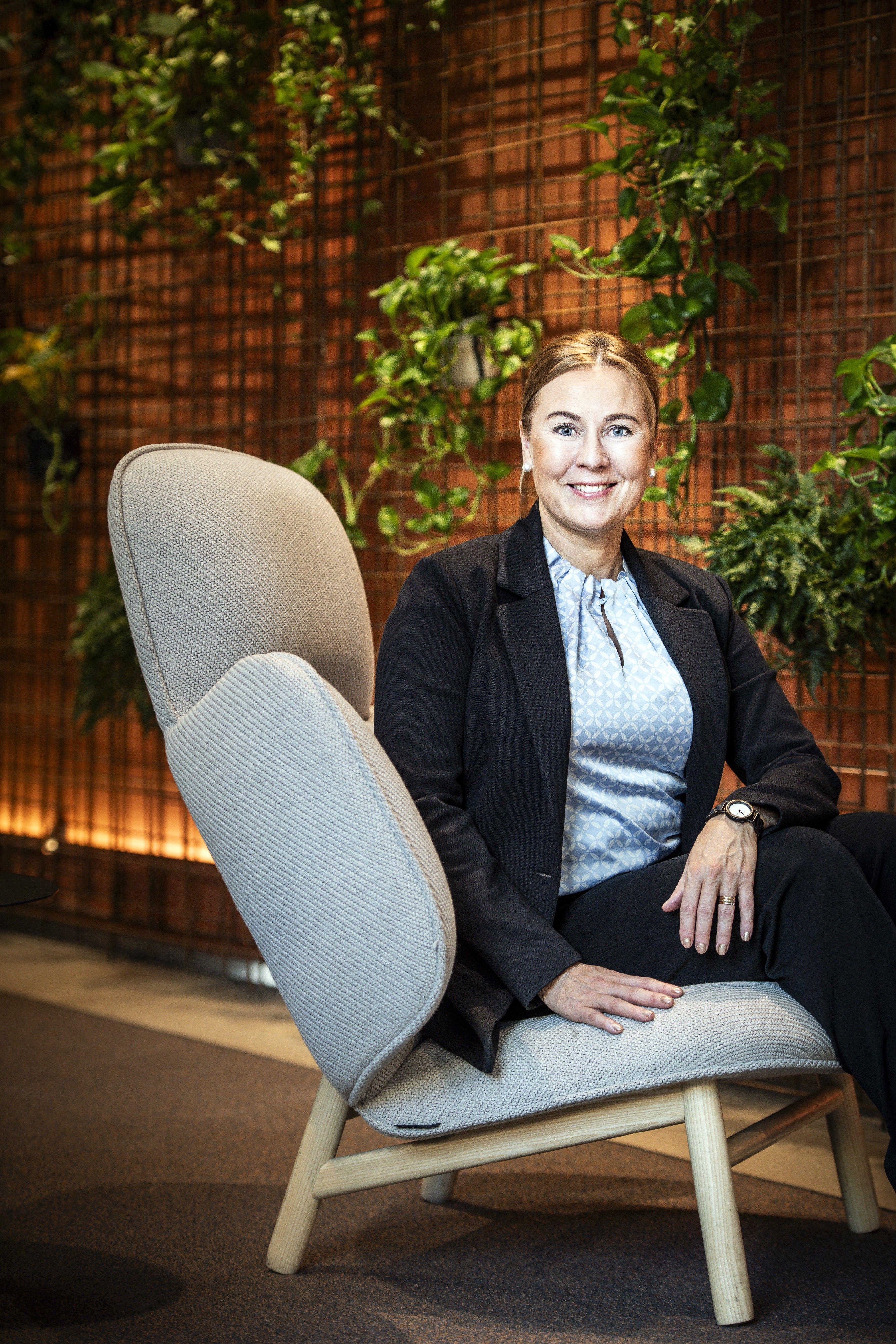 Harmaalla nojatuolilla istuva vaaleahiuksinen nainen katsoo suoraan kameraan ja hymyilee. Taustalla näkyy ruskeansävyiseen seinään kiinnitettyjä viherkasveja. Naisella on päällään vaaleanharmaa pusero sekä tummat housut ja jakku.
