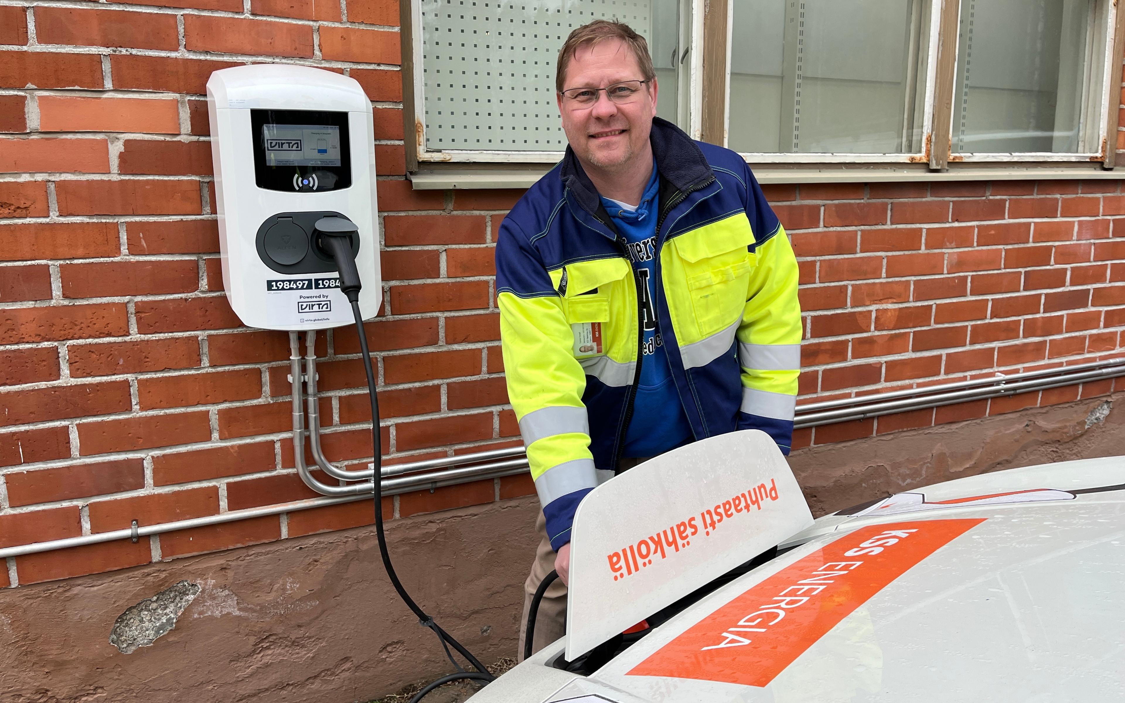 Hymyilevä henkilö huomiotakissa kiinnittää sähköautoa seinässä olevaan latauspisteeseen. Auton latausluukussa on teksti Puhtaasti sähköllä.