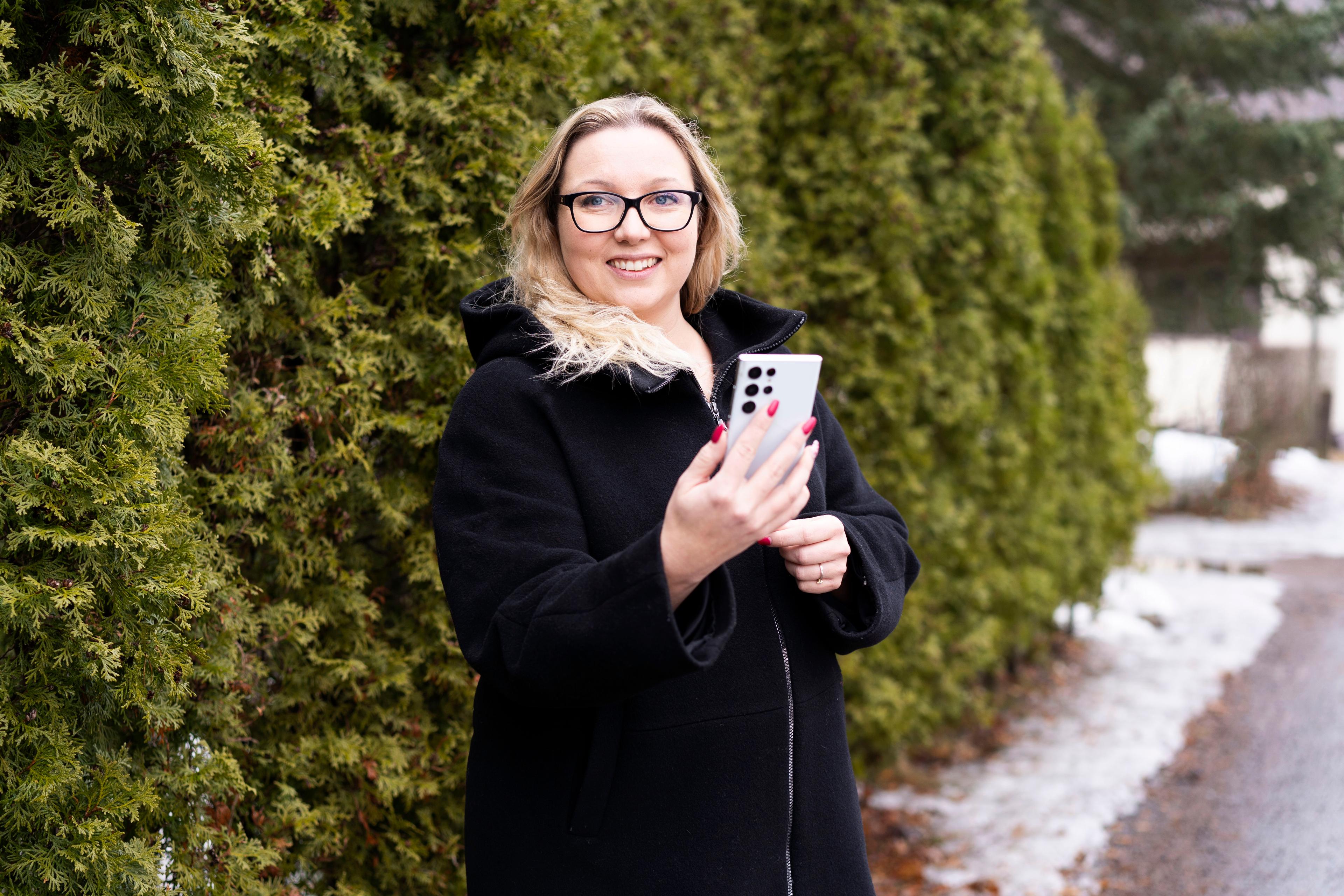 Mustaan talvitakkiin pukeutunut vaaleahiuksinen nainen hymyilee puhelin kädessään vihreän havuaidan edustalla. Naisella on pitkät punaiset kynnet ja valkoinen puhelin. Taustalla näkyy pihatietä, jonka varrella on hieman lunta.