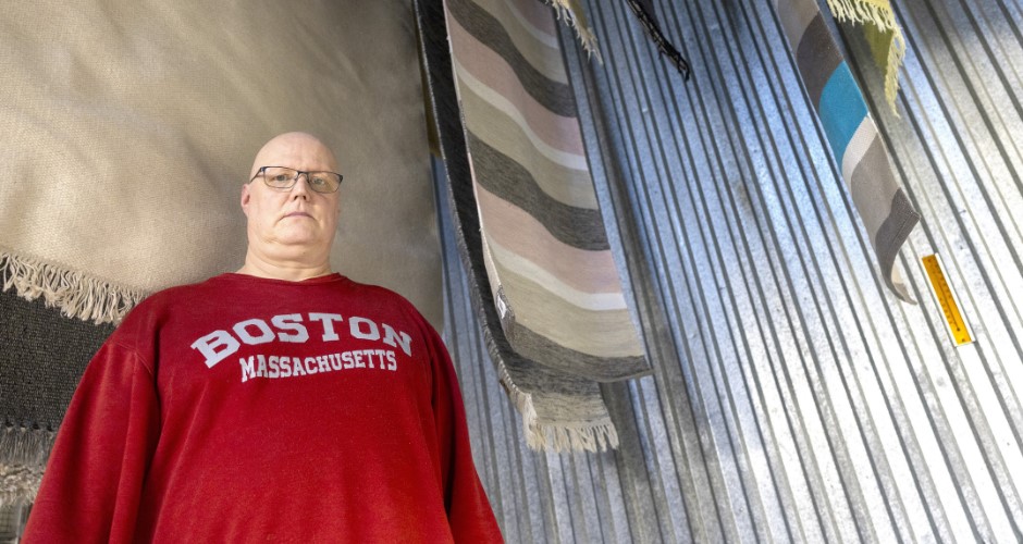 Punaiseen kollegepaitaan pukeutunut mies seisoo kuvan vasemmassa reunassa suoraan kameraan katsoen. Taustalla näkyy mattopesulan mattoja kuivumassa. Mattojen taustalla on aaltoileva harmaa peltiseinä.