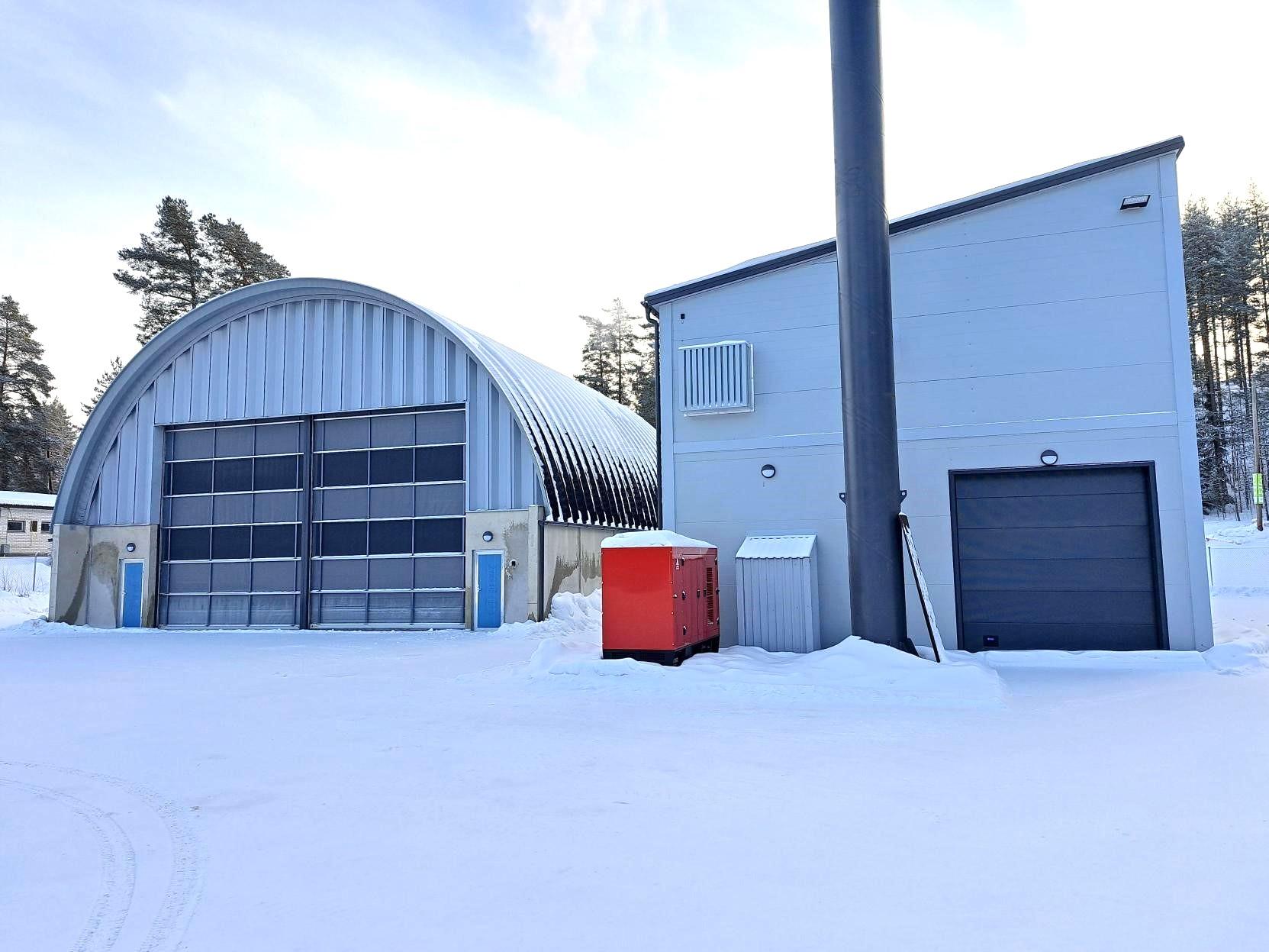 Kuvassa kaksi harmaata metallista rakennusta. Oikeanpuoleinen rakennus on biolämpölaitos, jonka edessä näkyy tumman harmaa piippu ja nosto-ovi. Vasemmanpuoleinen kaarihalli on polttoainesiilo.
