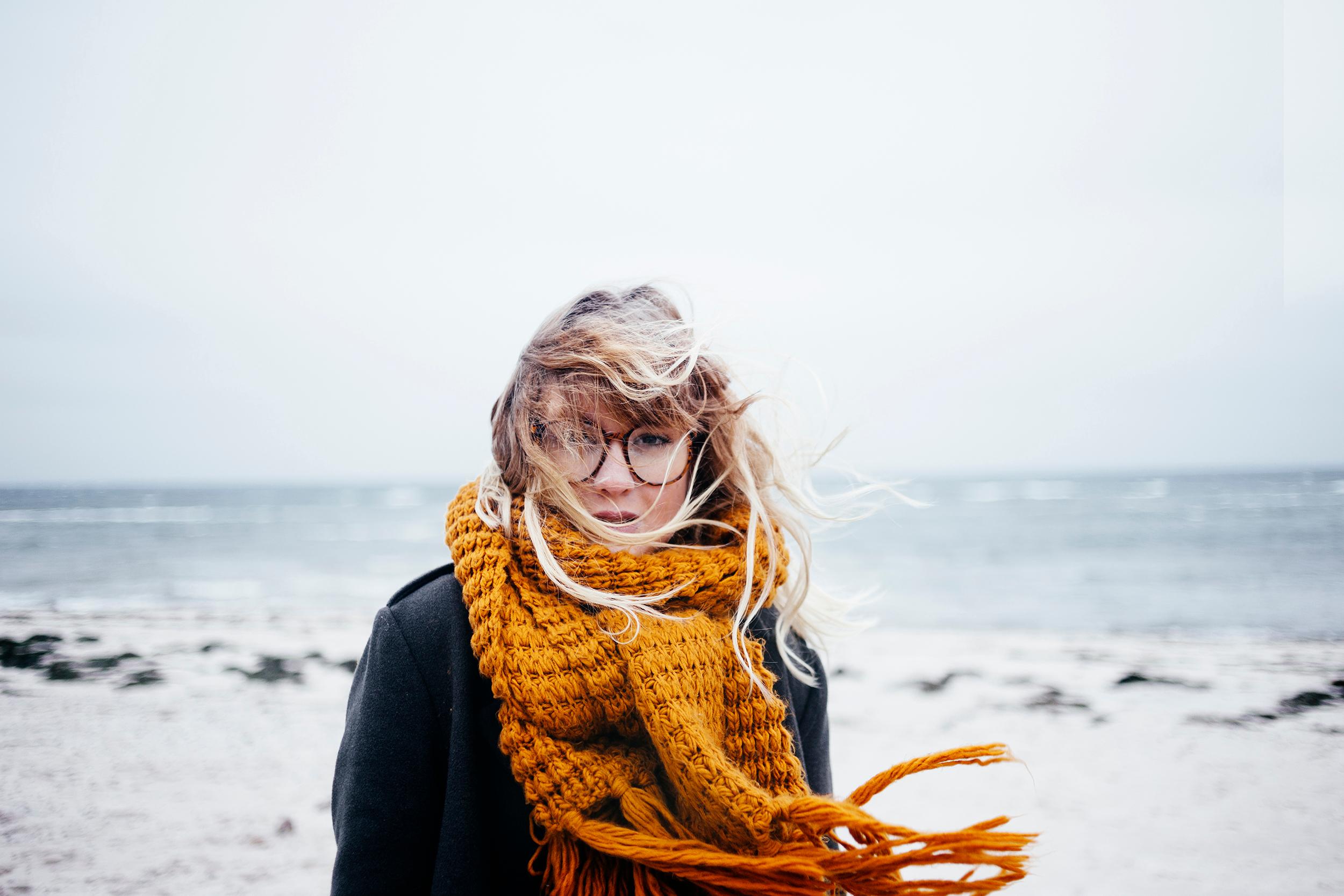 Nainen seisoo tuulisella rannalla syksyisessä säässä. Hänellä on musta takki, muhkea oranssi kaulaliina ja pyöreät silmälasit. Hiukset ja kaulaliina liikkuvat tuulen mukana.