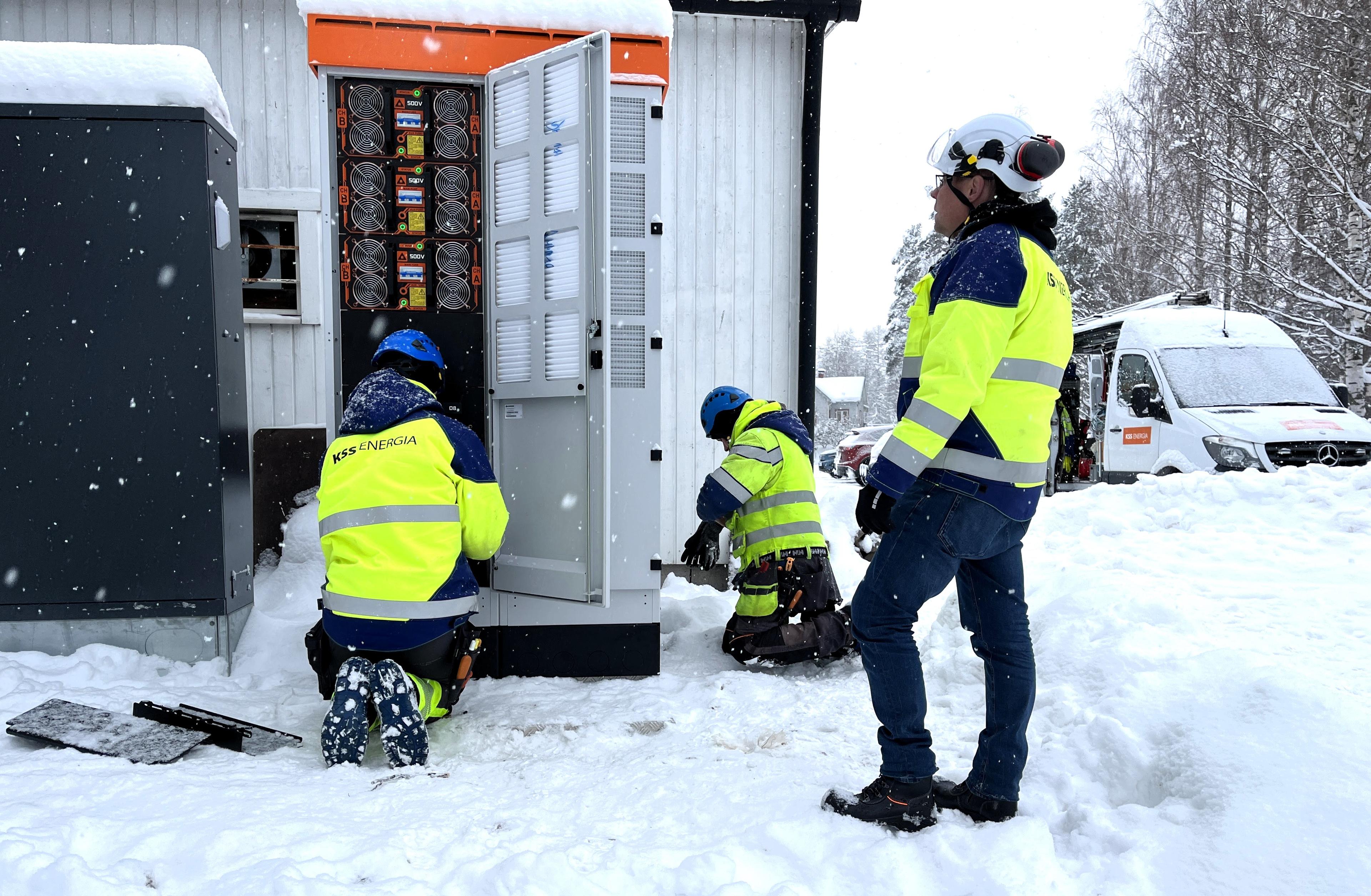 Kolme asentajaa työskentelee lumisissa maisemissa rakennuksen sähkökaapin parissa. 