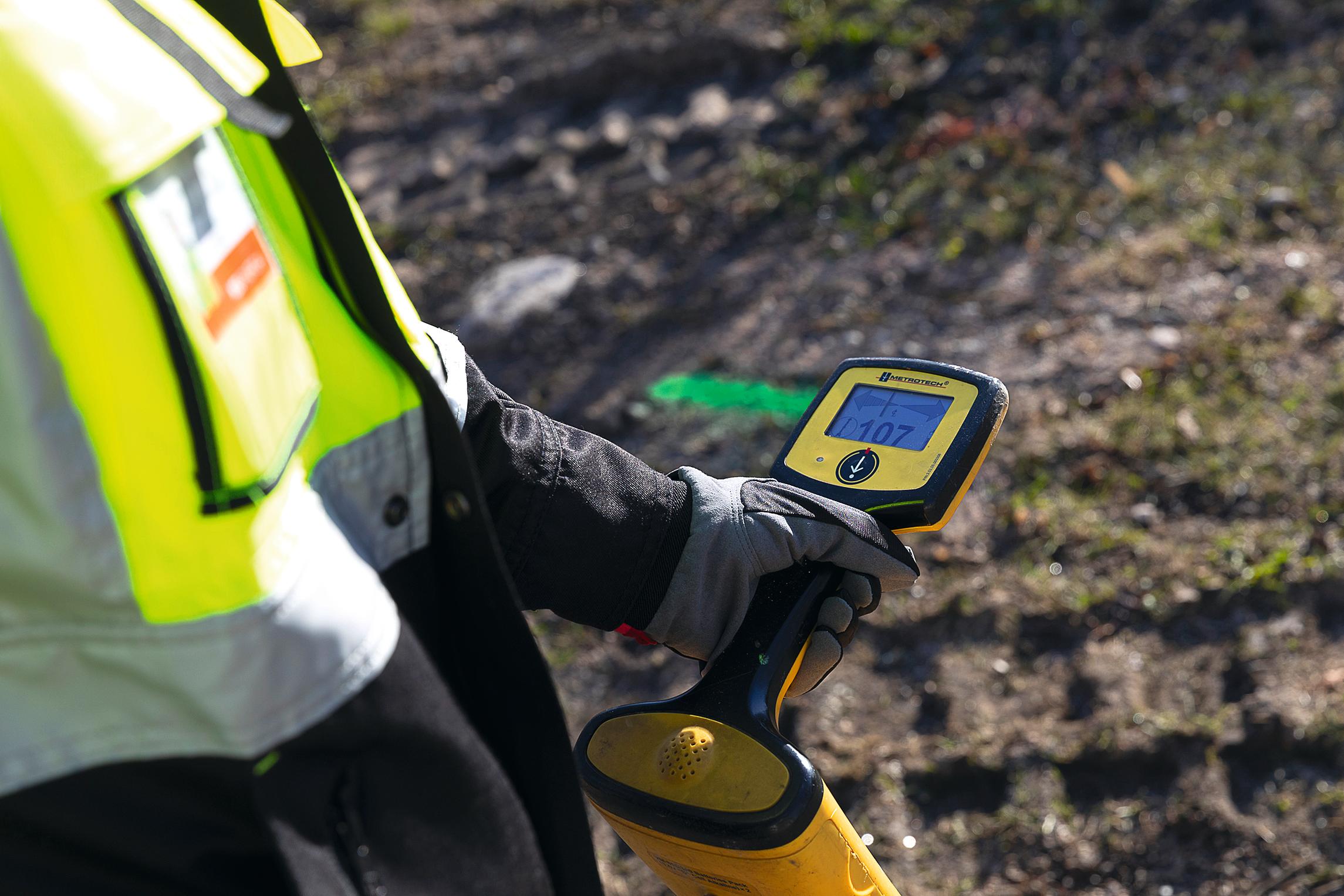 Lähikuva henkilön kädessä olevasta keltaisesta mittauslaitteesta, jolla tutkitaan maassa olevien kaapeleiden sijaintia.