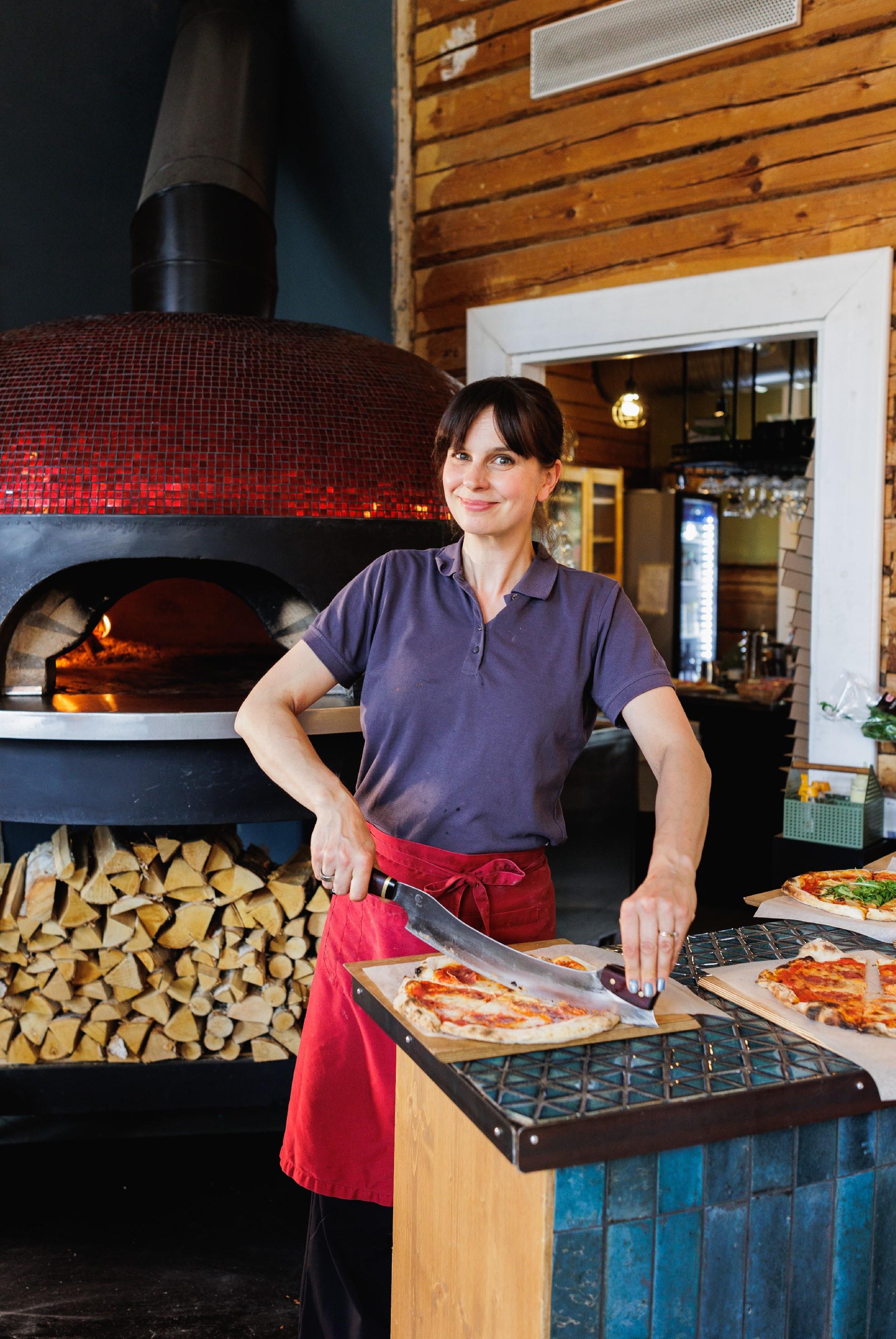 Näyttelijä-ravintoloitsija Olga Temonen leikkaa pizzaa suoraan kameraan katsoen Ravintola Rehtorin suuren punaisen pizzauunin edustalla.