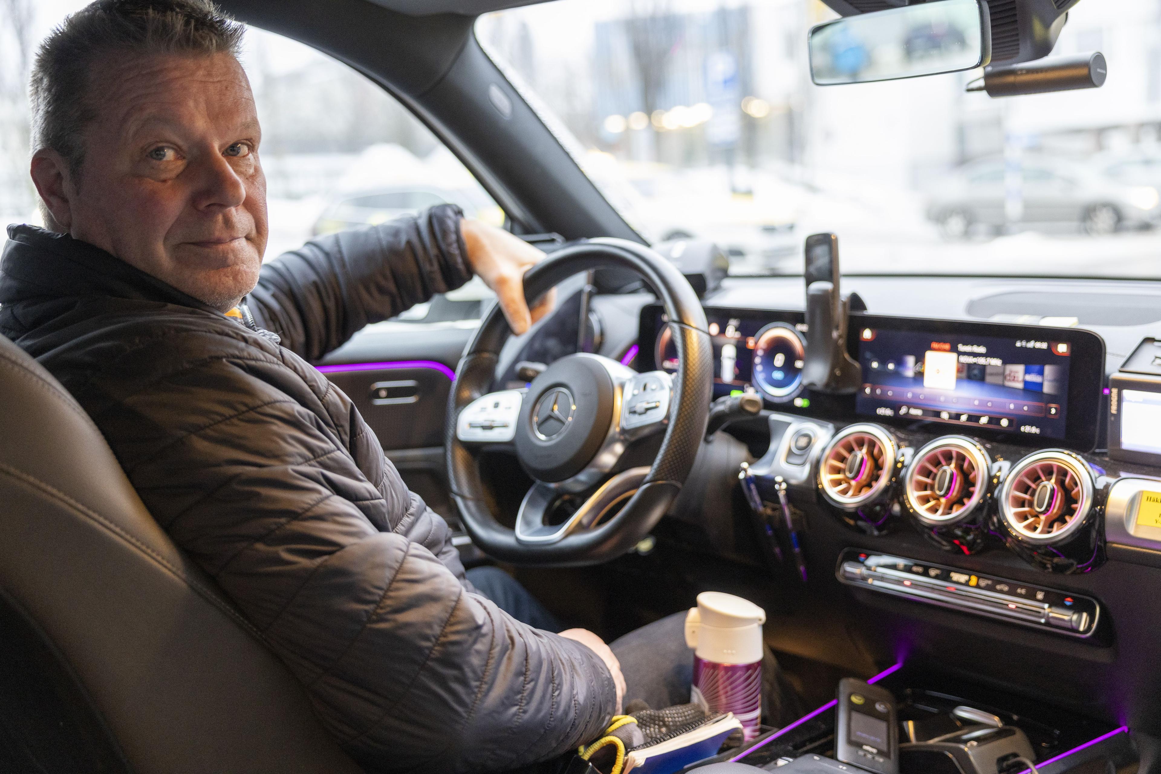 Tummatakkinen mies istuu autossa kuljettajan paikalla, vasen käsi auton ratissa kiinni. Kuva on kuvattu auton sisällä takapenkin suunnasta etupenkkiä kohti.