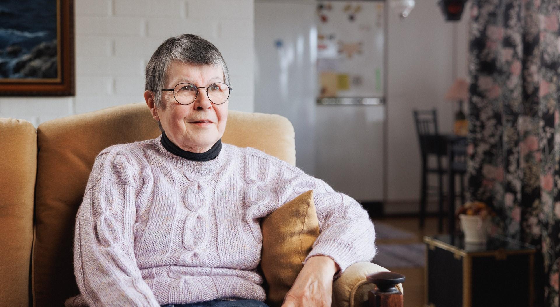 Vaaleaan neuleeseen pukeutunut lyhythiuksinen nainen istuu sohvalla haastattelutilanteessa. Eläkeikäisellä naisella on silmälasit ja hän hymyilee hieman. Taustalla näkyy valkoinen tiiliseinä ja taulu, hieman kauempana keittiö.