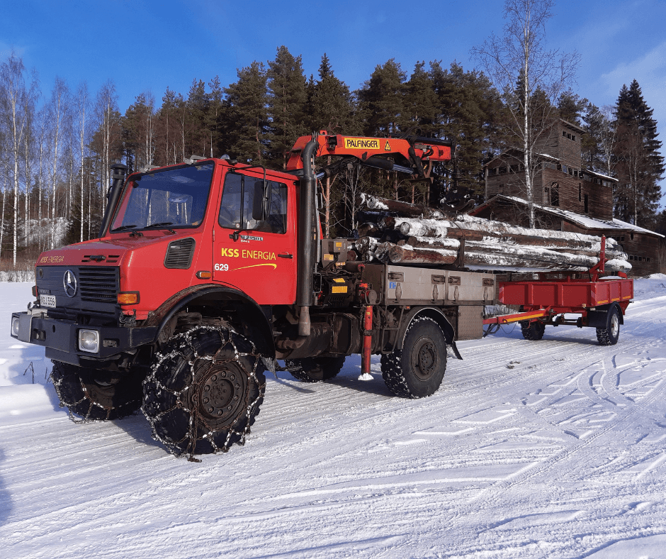 Punainen maastokuorma-auto aurinkoisena talvipäivänä. Maastokuorma-auton kyydissä lumisia sähköpylväitä, taustalla näkyy havu- ja koivumetsää.