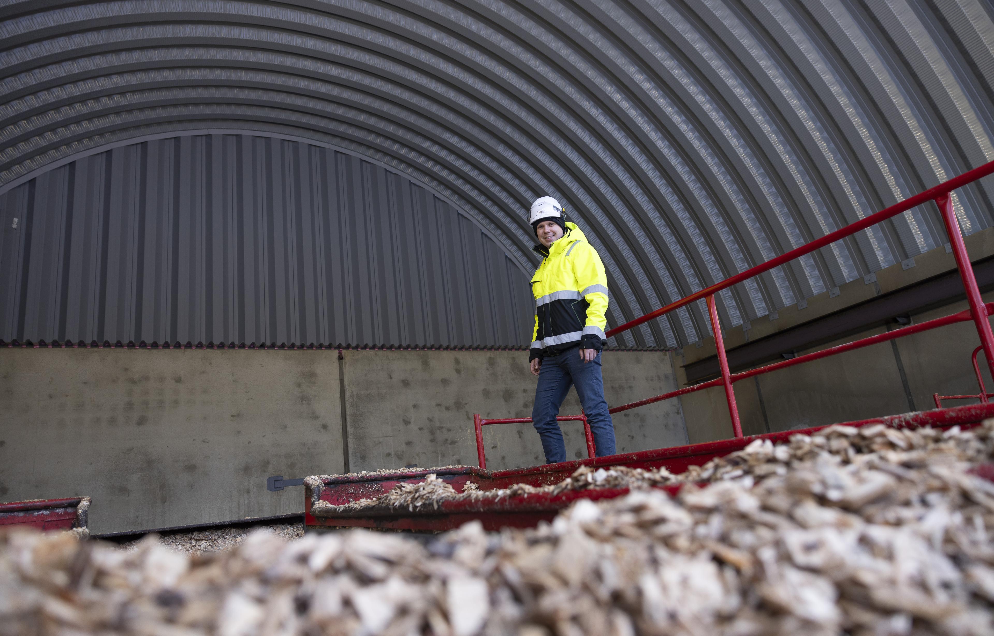 Kirkkaan keltaiseen työmaatakkiin ja valkoiseen kypärään pukeutunut mies seisoo hakesiilon edustalla. Taustalla näkyy kaareva halli, edessä puuhaketta. Mies katsoo suoraan kameraan ja seisoo punakaiteisella ritiläkäytävällä.