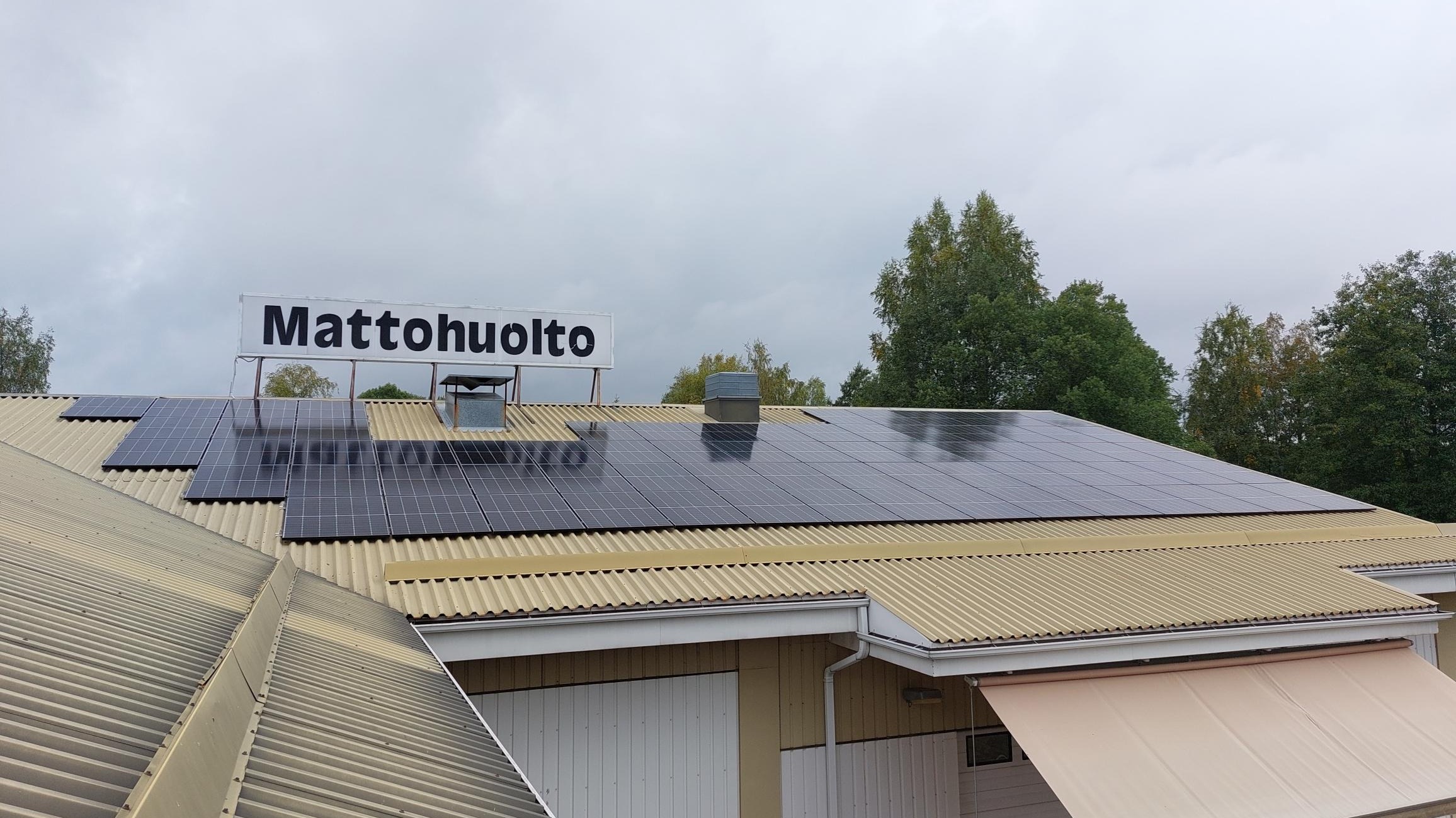 Kattokuva keltaisesta rakennuksesta, jonne on asennettu katolle tummat aurinkopaneelit. Katolla näkyy valkoinen kyltti, jossa lukee mustalla tekstillä Mattohuolto. Rakennuksen takana on vehreiden puiden latvoja, sää on pilvinen.
