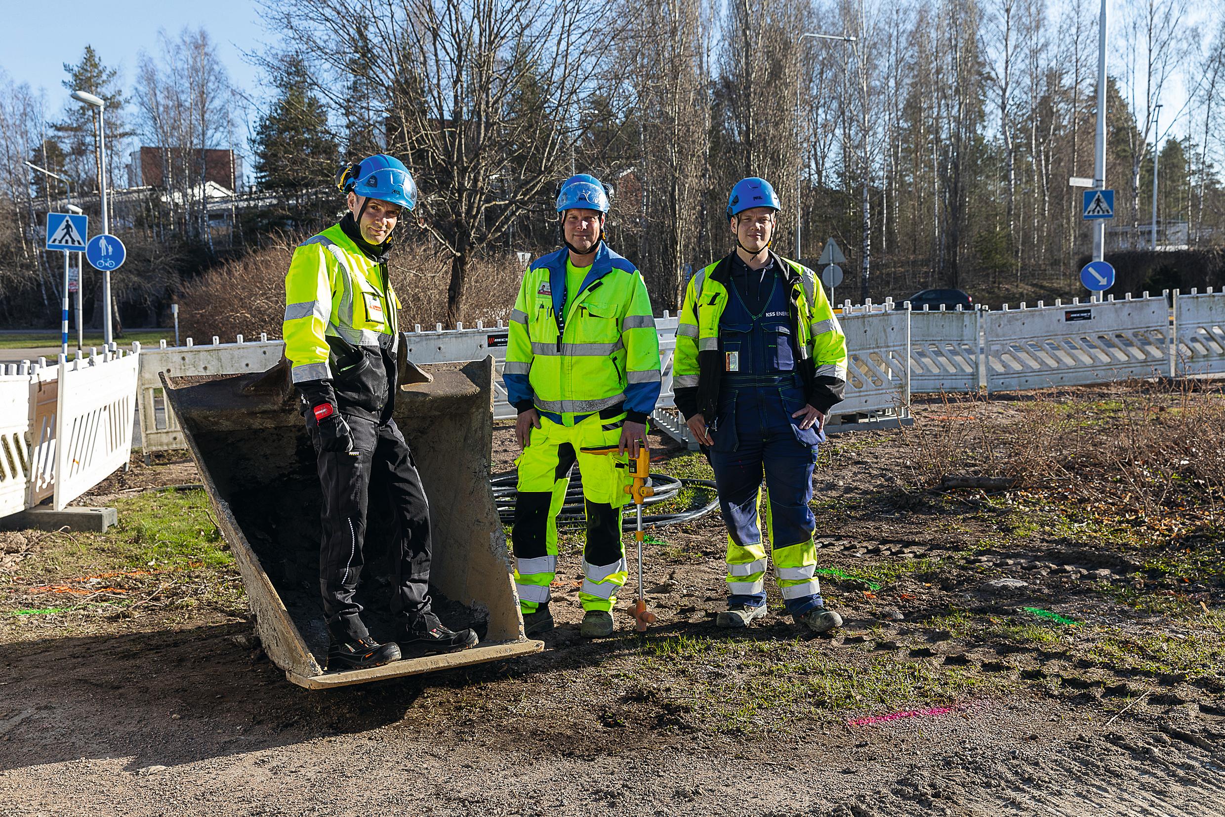 Kolme henkilöä suojavarusteissa, yksi heistä kaivurista irrotetun kauhan sisällä. Heillä on kaapelinetsinnässä ja -merkinnässä tarvittavia työvälineitä.