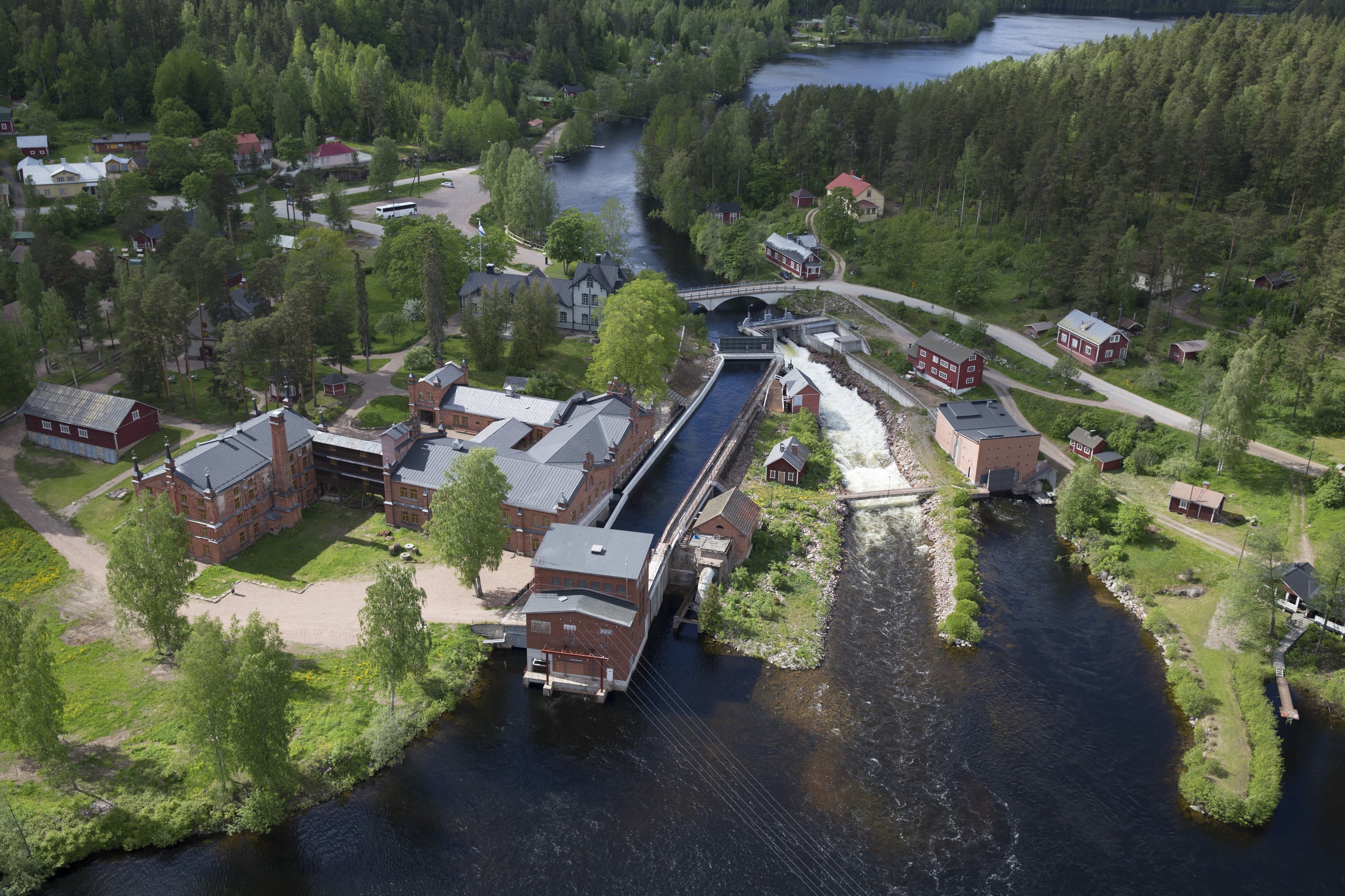 Historialliset punatiiliset puuhiomorakennukset vasemmalla ja keskellä voimalaitosrakenteet koskessa. Pato on auki ja koski kuohuu. Ympärillä näkyy metsää ja taloja sekä vesistöä.