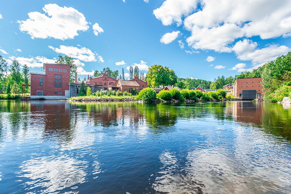 Kuvassa näkymä väreilevän veden yli Verlan vesivoimalaitokselle. Kuvassa näkyy veden takana taustalla punatiilisiä rakennuksia, puita, pensaita ja kirkas taivas, jossa valkeita pilvenhattaroita.