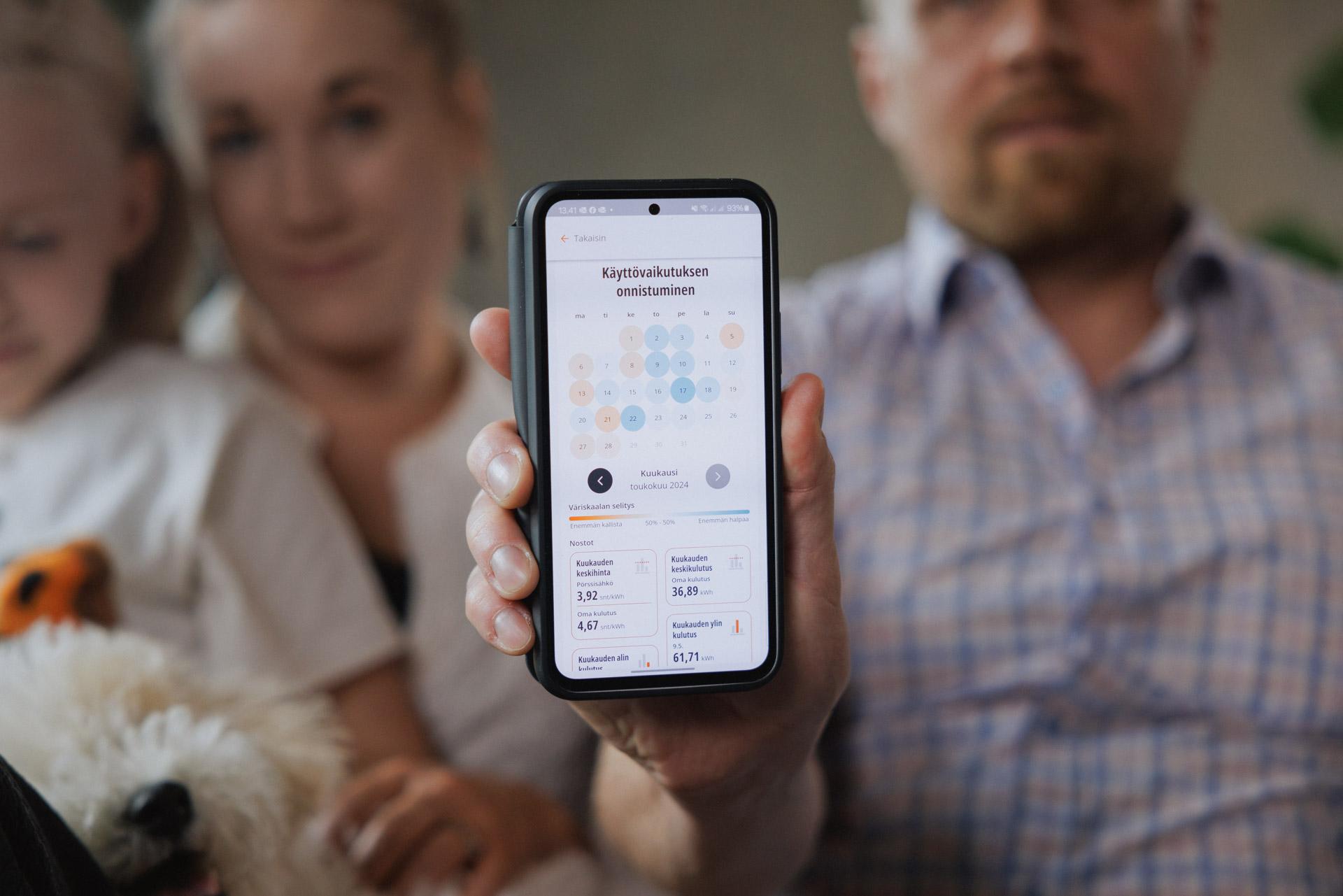 Lähikuva kännykän ruudusta. Puhelin on ojennettu kohti kameraa ruutupaitaisen miehen toimesta. Hänen vierellään istuu nainen, jonka sylissä on kouluikäinen tyttö sekä valkea pieni koira.