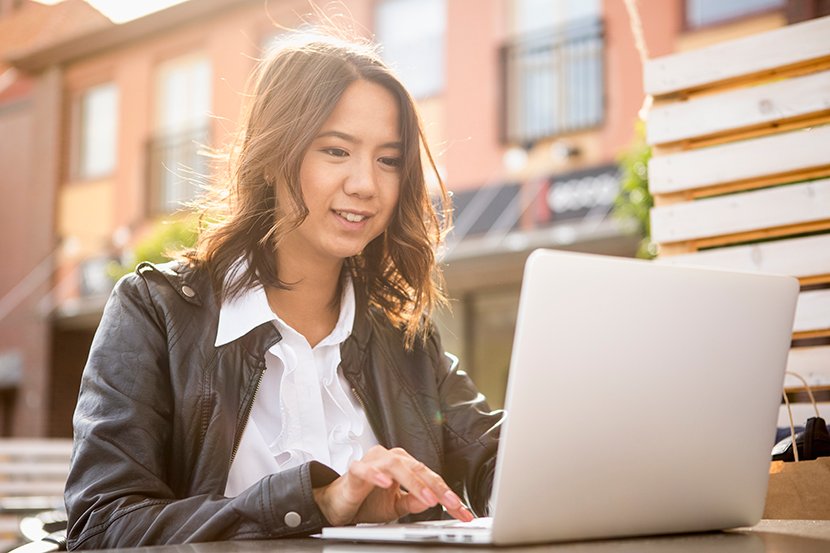 Tummahiuksinen hymyilevä nainen istuu harmaan kannettavan tietokoneen ääressä ulkona. Naisella on päällä valkoinen kauluspaita ja ruskea nahkatakki. Taustalla näkyy vaaleanpunainen kerrostalo.