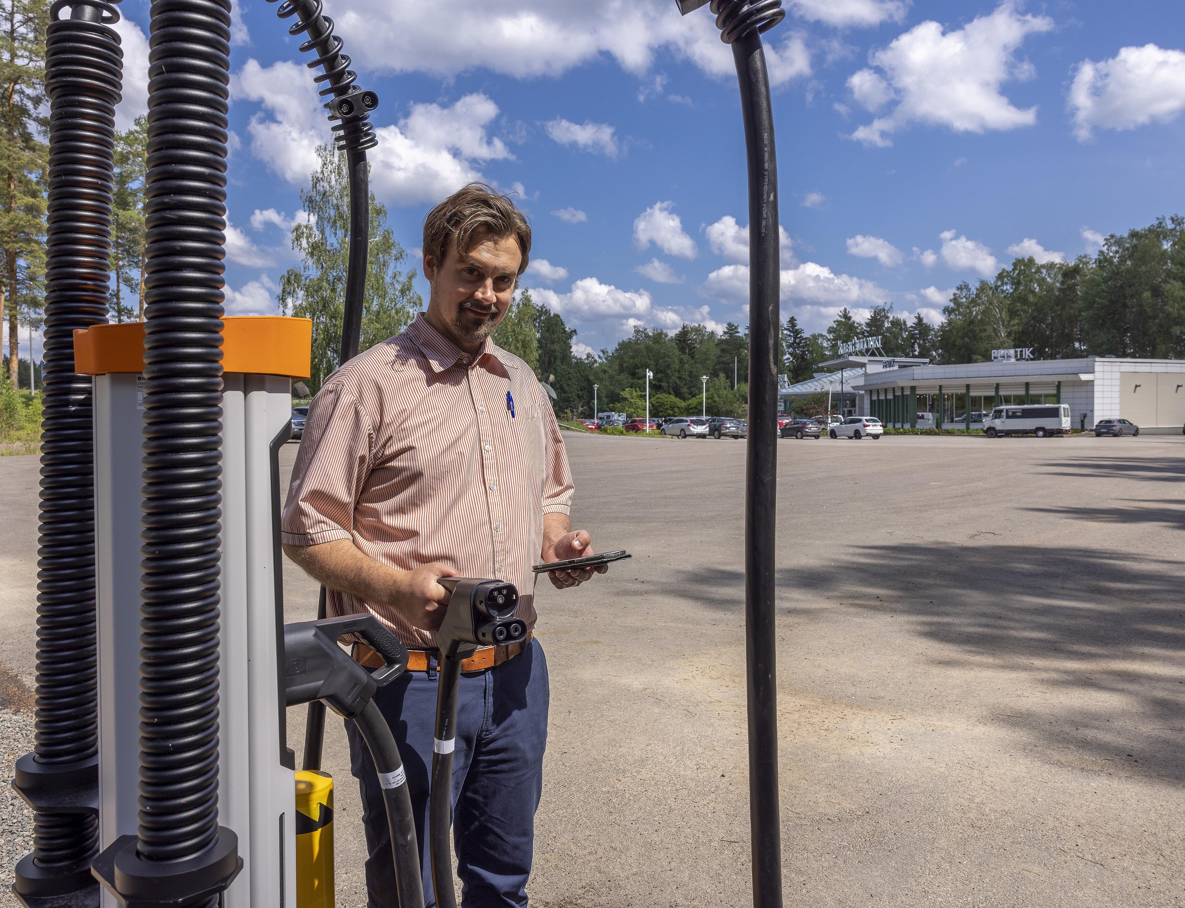 Kymen Paviljongin yrittäjä Lauri Leskinen sähköauton suurteholaturi kädessään ja suoraan kameraan katsoen. Takana näkyy Kymen Paviljonki ja kesäisen sininen taivas poutapilvillä.