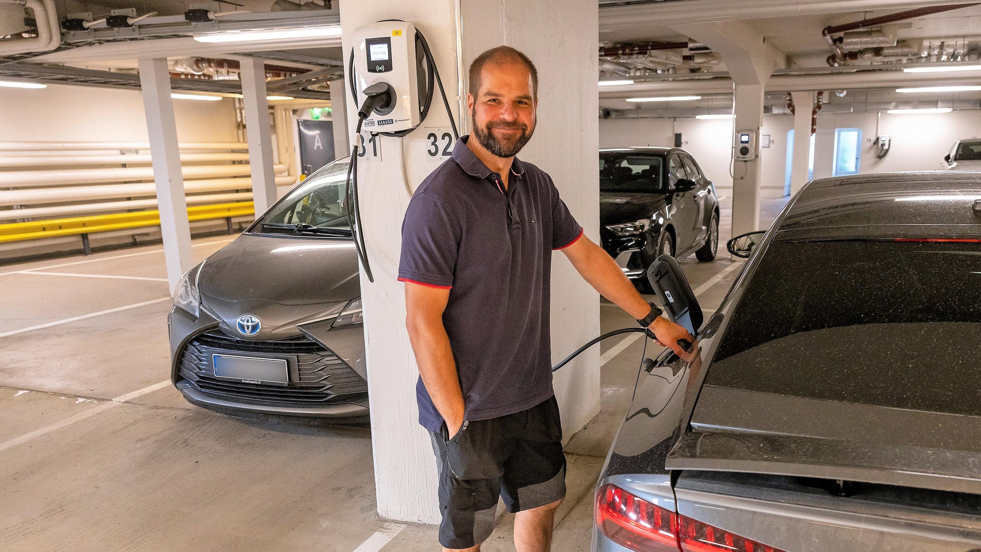 As Oy Helsingin Lauttasaarenrannan hallituksen puheenjohtaja Jukka Vakula lataa harmaata sähköautoaan hymyillen ja suoraan kameraan katsoen.