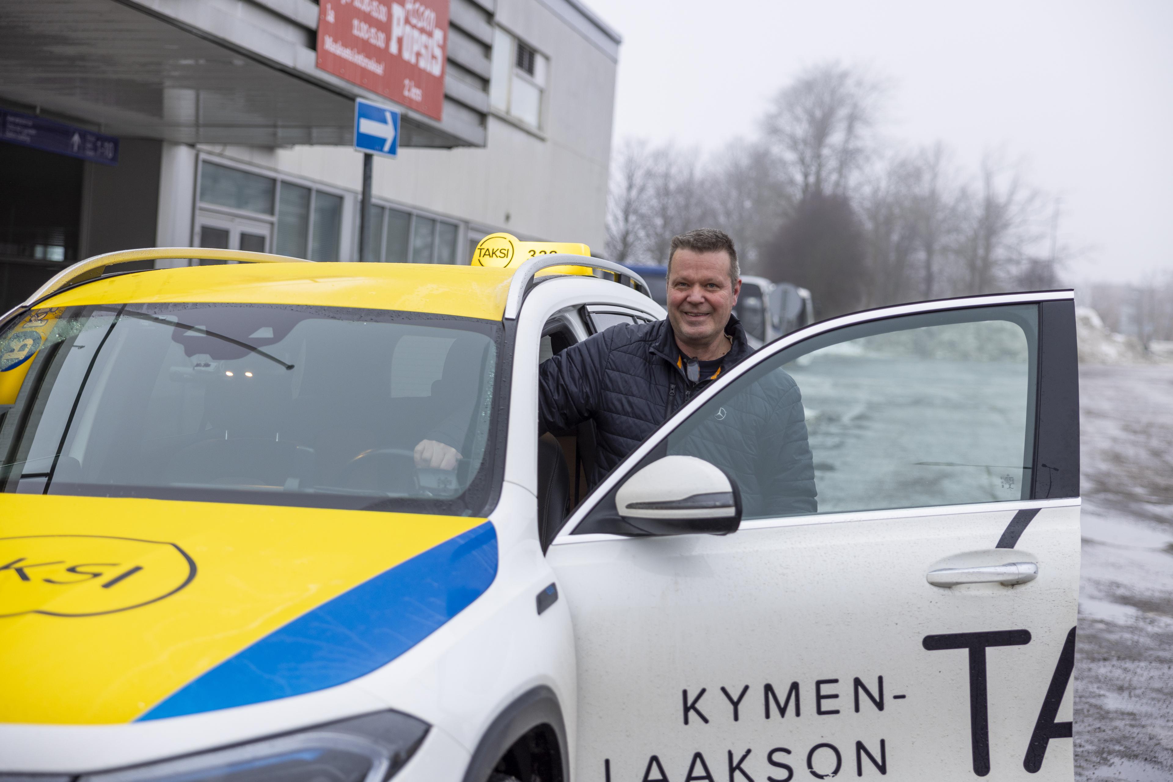 Taksiautoilijamies seisoo kuljettajan puolen oven luona auki olevan oven takana ja hymyilee. Miehellä on musta talvitakki ja lyhyet hiukset. Auto on teipattu valkoisen, keltaisen ja sinisen sävyin. Taustalla näkyy harmaa rakennus, linja-autoja ja puita. Ilma on alkutalven harmaa.