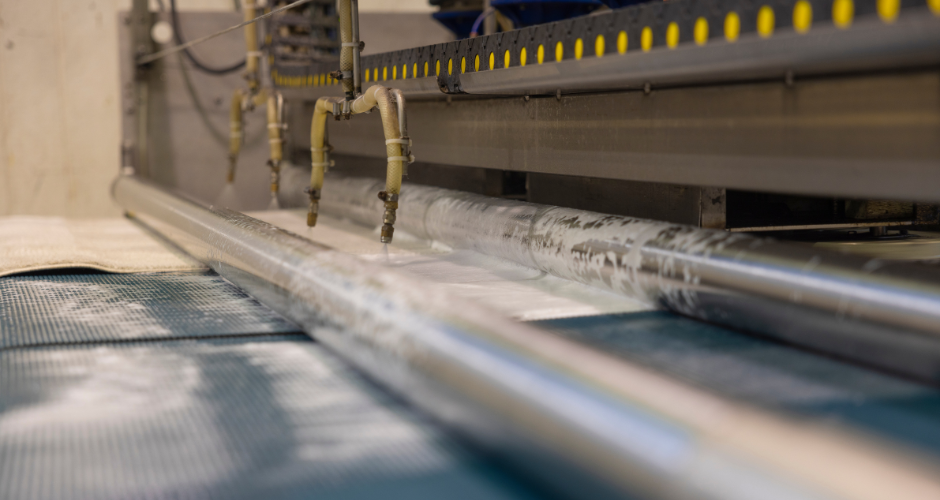 Lähikuva matonpesukoneesta. Ruskea-harmaan väritteisessä kuvassa näkyy koneen mattoa ohjaavia pitkiä rullia. Kuvan ylälaidassa näkyy koneeseen liimattua keltaista varoitusnauhaa.