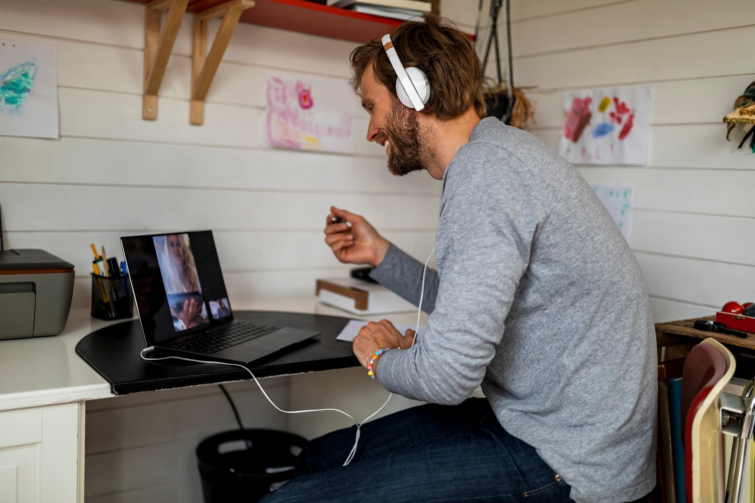 Mies istuu harmaassa paidassa ja kuulokkeet päässä työhuoneessa, jossa on seinillä lasten värikkäitä piirrustuksia ja katsoo tietokoneen ruutua, jossa näkyy toisen henkilön kasvot.
