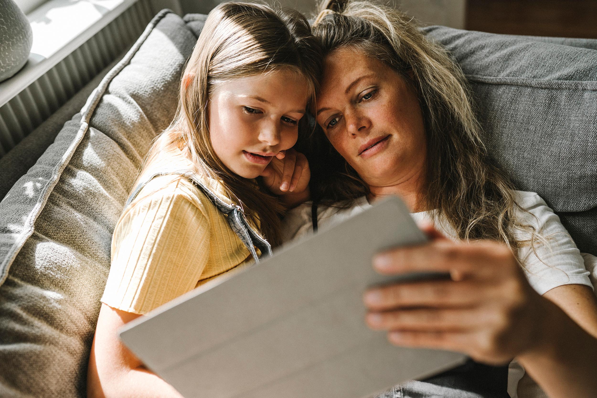 Tyttö ja äiti katsovat keskittyneesti tablettia yhdessä sylikkäin maaten vaalean harmaalla sohvalla auringon paistaessa ikkunasta heidän kasvoille. Molemmilla on pitkät hiukset. Tytöllä on vaaleankeltainen paita ja farkkuhaalarit ja äidillä valkoinen t-paita. 