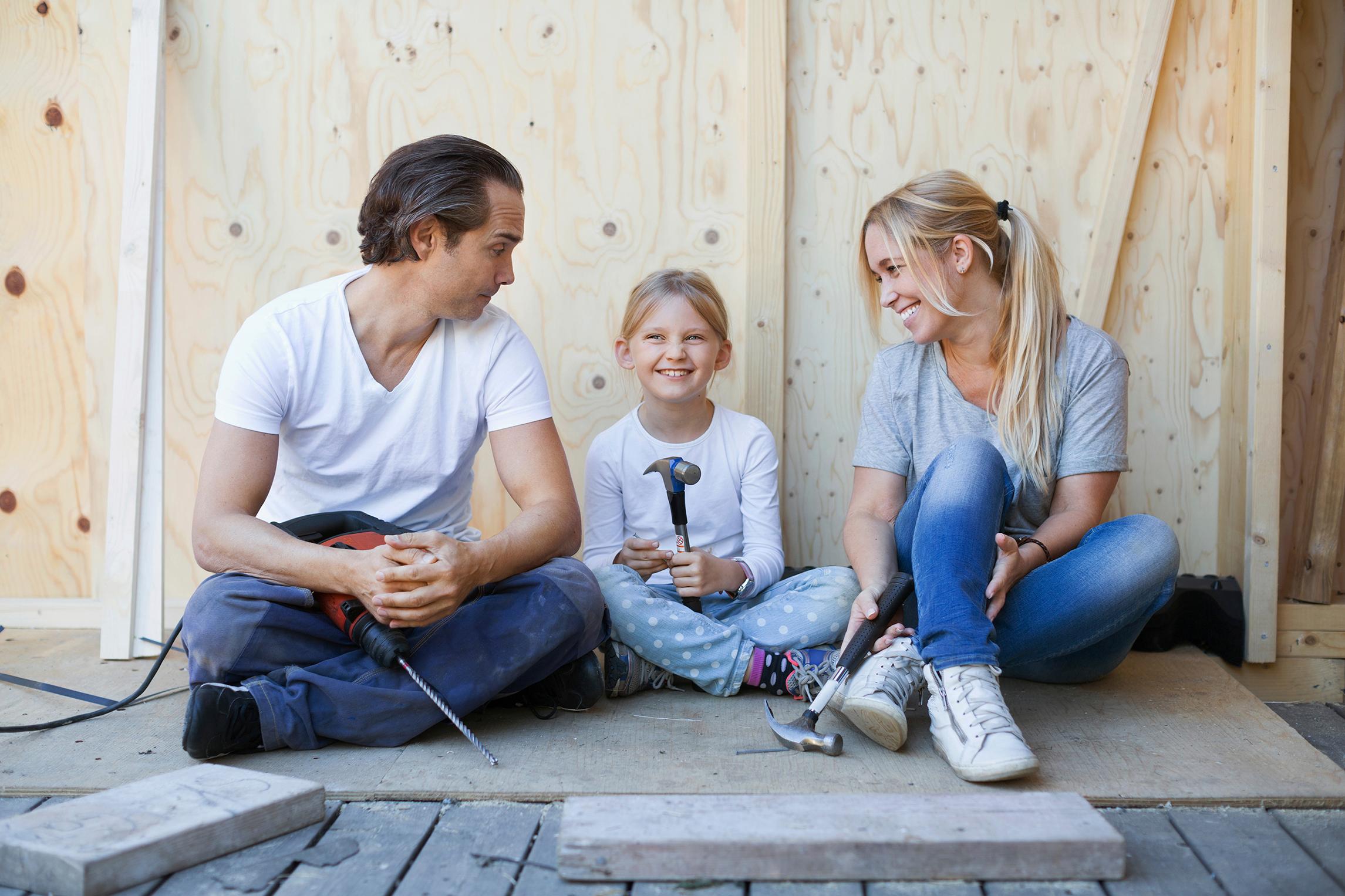 Isä, äiti ja lapsi istuvat rakennustyömaalla työkalut käsissään.