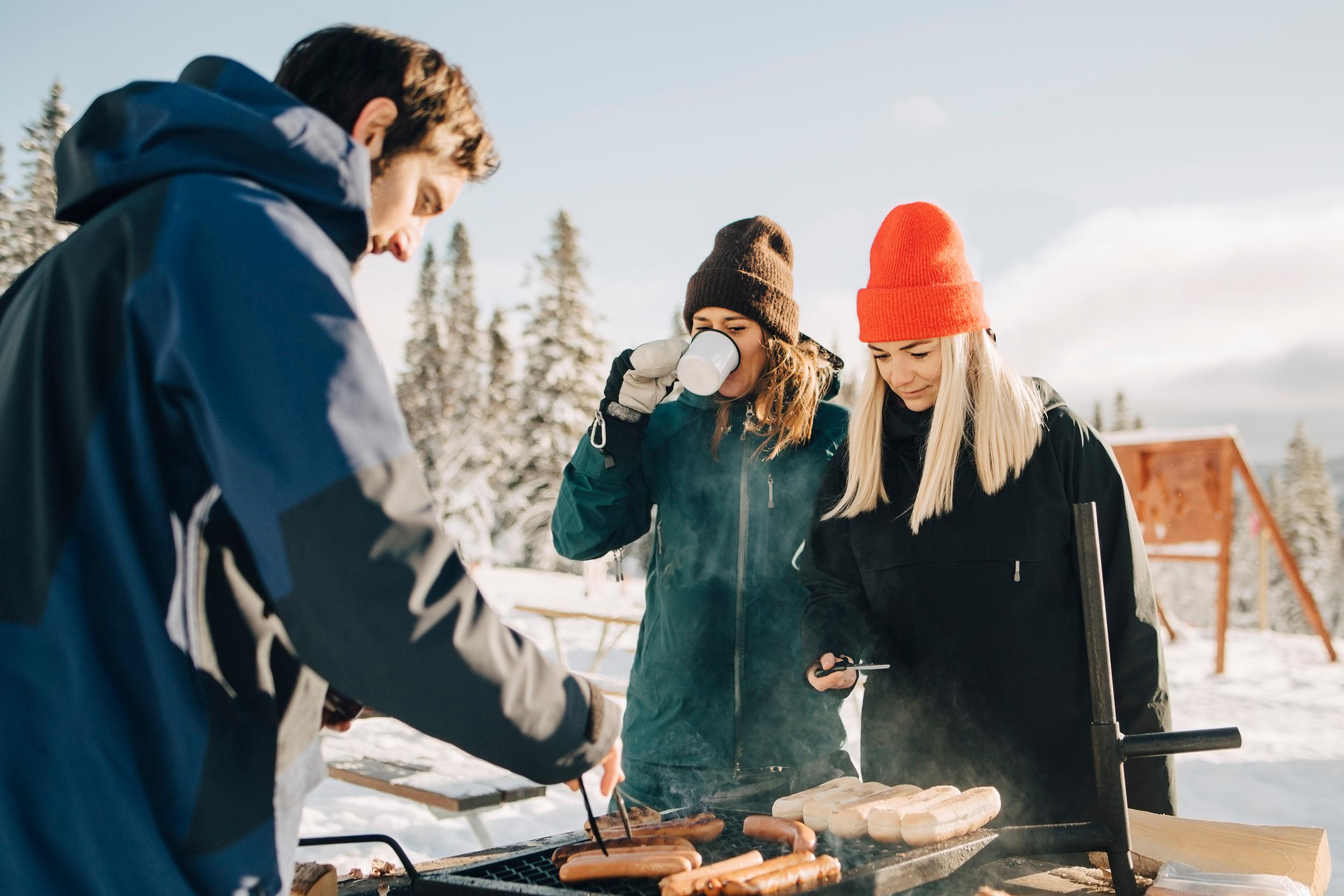 Kolme nuorta aikuista grillaavat makkaraa talvisessa säässä. Kuvan etualalla sinimustaan toppatakkiin pukeutunut ruskeahiuksinen mies kääntelee pihdeillä makkaroita selin kameraan. Kaksi pipopäistä ja pitkähiuksista naista ovat kasvot kameraan päin, toinen naisista juo valkoisesta mukista. Taustalla näkyy lumista kuusimetsää ja pilvinen taivas.