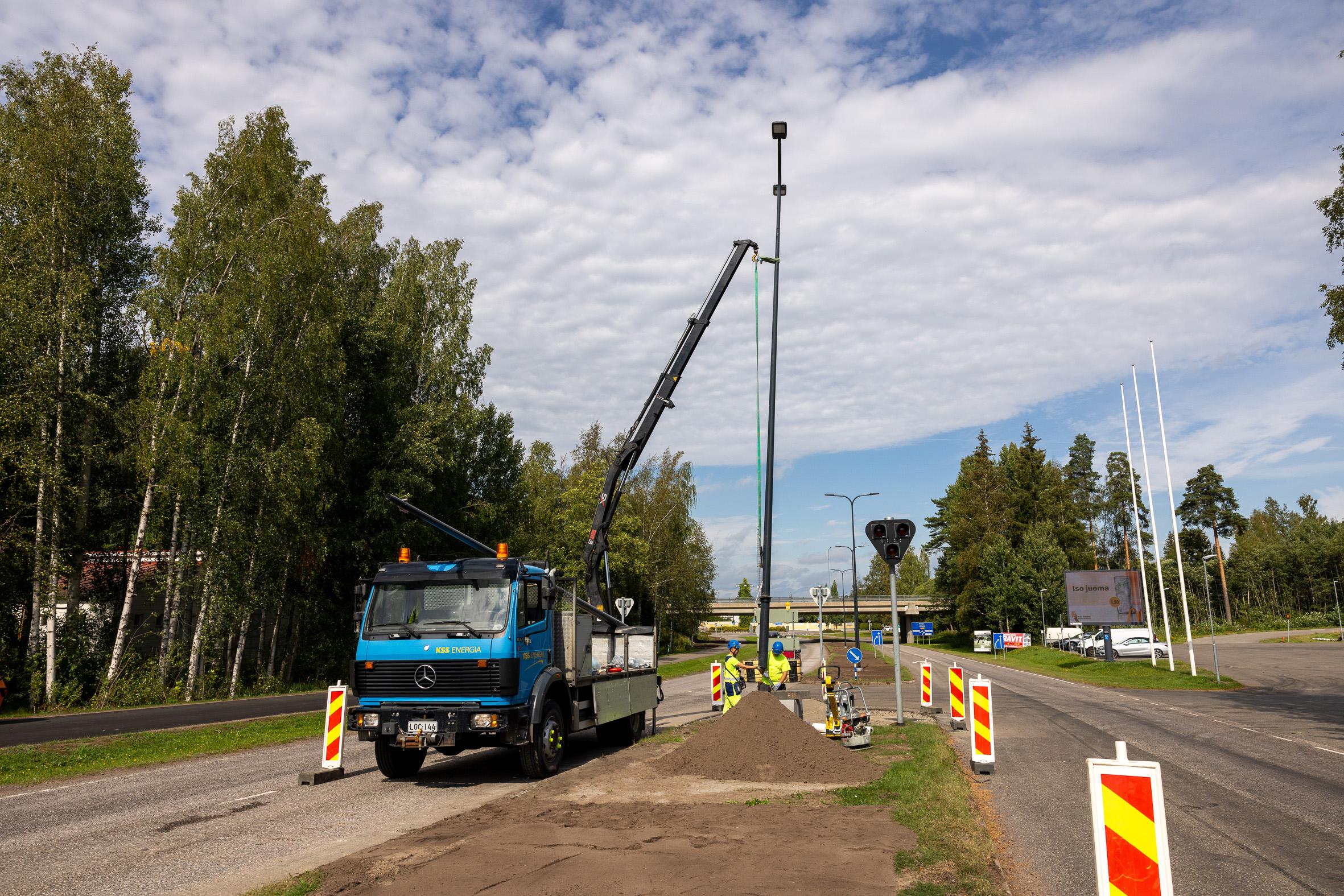 Nosturilla nostetaan katuvalopylvästä pystyyn. Kaksi henkilöä suojavarusteissaan ohjaavat nostoa.