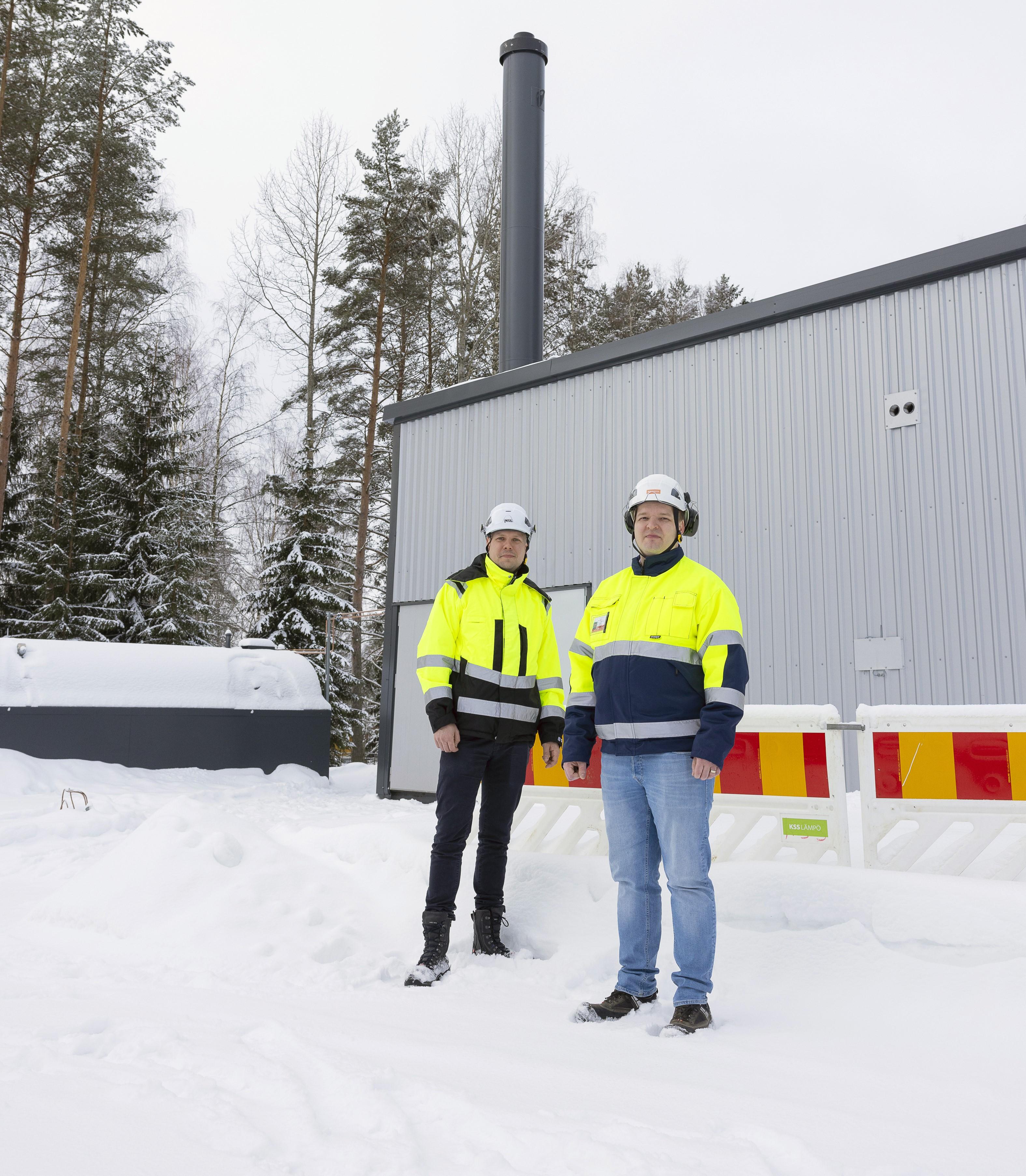 Kaksi kirkkaan keltaisella värillä varustettuihin työtakkeihin ja valkoisiin kypäröihin pukeutunutta miestä seisoo harmaan lämpölaitoksen edustalla. Talvisessa maisemassa näkyy taustalla puista. Miesten taustalla on keltapunainen työmaa-aita.