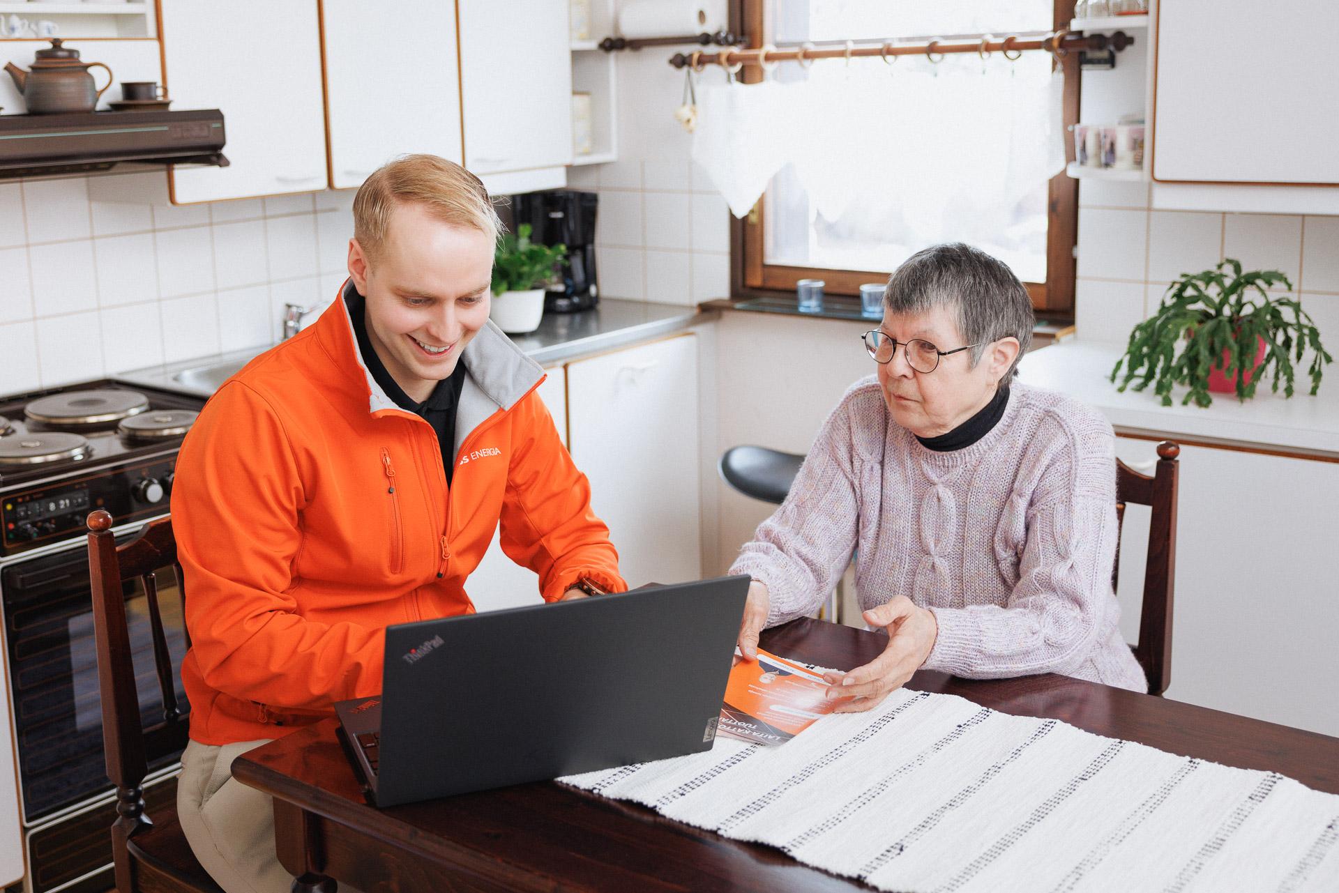 Oranssiin takkiin pukeutunut mies ja vaaleanviolettiin neuleeseen pukeutunut nainen katsovat keittiönpöydän äärellä. Mies hymyilee ja esittelee vanhemmalle rouvalle kannettavan tietokoneen näyttöä. Molemmat katsovat näytölle. Takana näkyy vaaleita keittiön kaappeja, hella sekä viherkasveja.