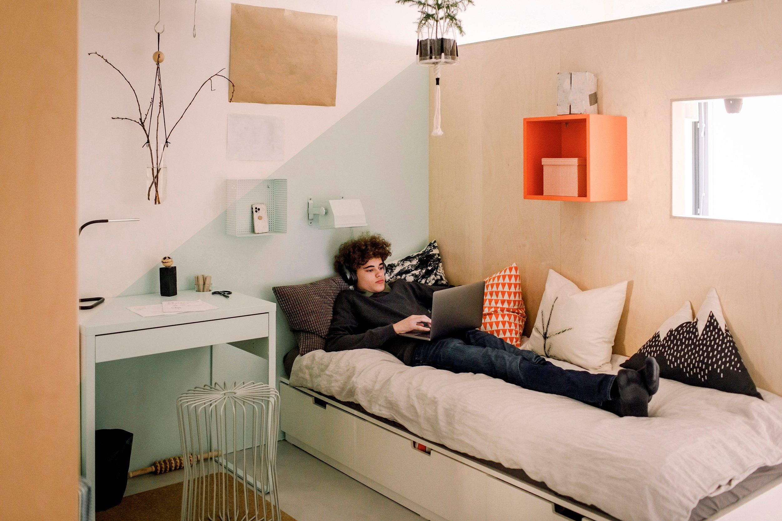 Nuori makaa sängyllä valkoisen sänkypeiton päällä ja katsoo kannettavaa tietokonetta kuulokkeet korvilla. Seinällä on oranssi hylly, jossa laatikko sisällä ja sängyn vieressä on yöpöytä, jossa lamppu ja muuta pientä tavaraa.