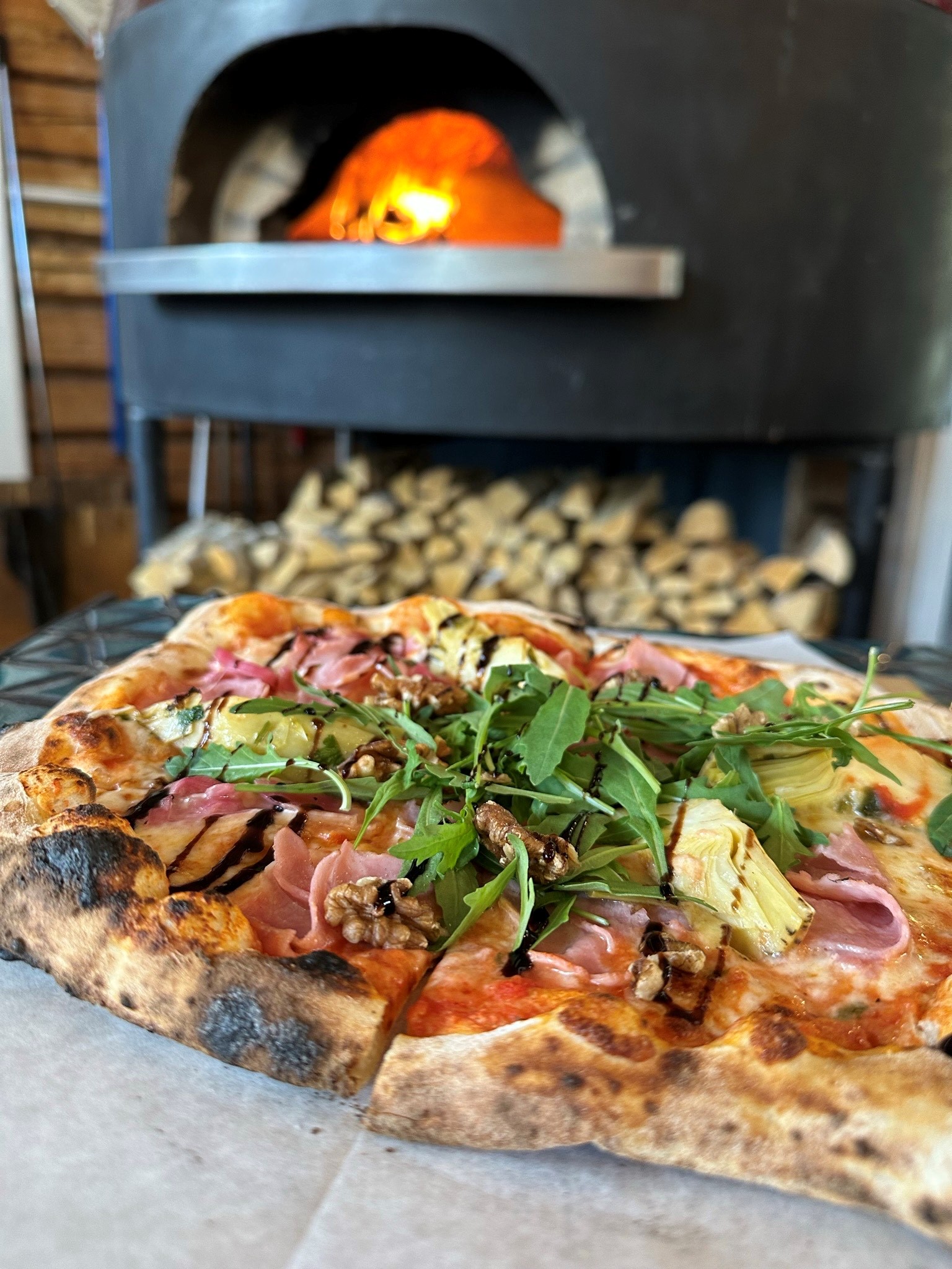 Lähikuva herkullisesta pizzasta, jossa täytteenä muun muassa uunikinkkua, marinoitua artisokkaa, saksanpähkinöitä ja päällimmäisenä vihreää rucolaa. Takana näkyy Ravintola Rehtorin suuri pizzauuni, jossa on tuli.